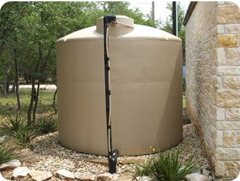 A beige plastic rainwater tank with a black downspout connected on top, situated outside near a stone wall with trees and plants in the background.