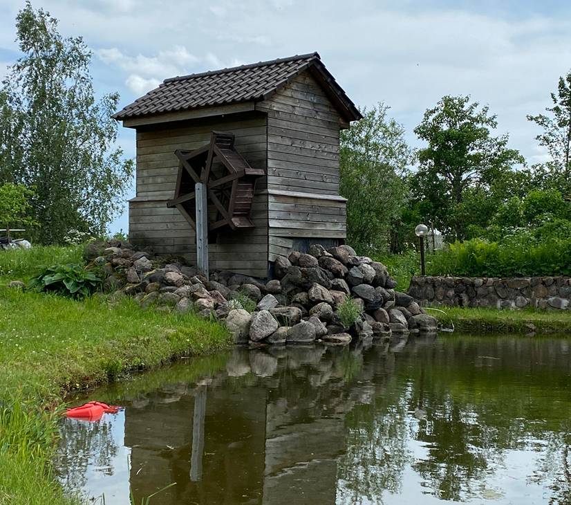 A wooden watermill with a water wheel located beside a pond, surrounded by trees and rocks.
