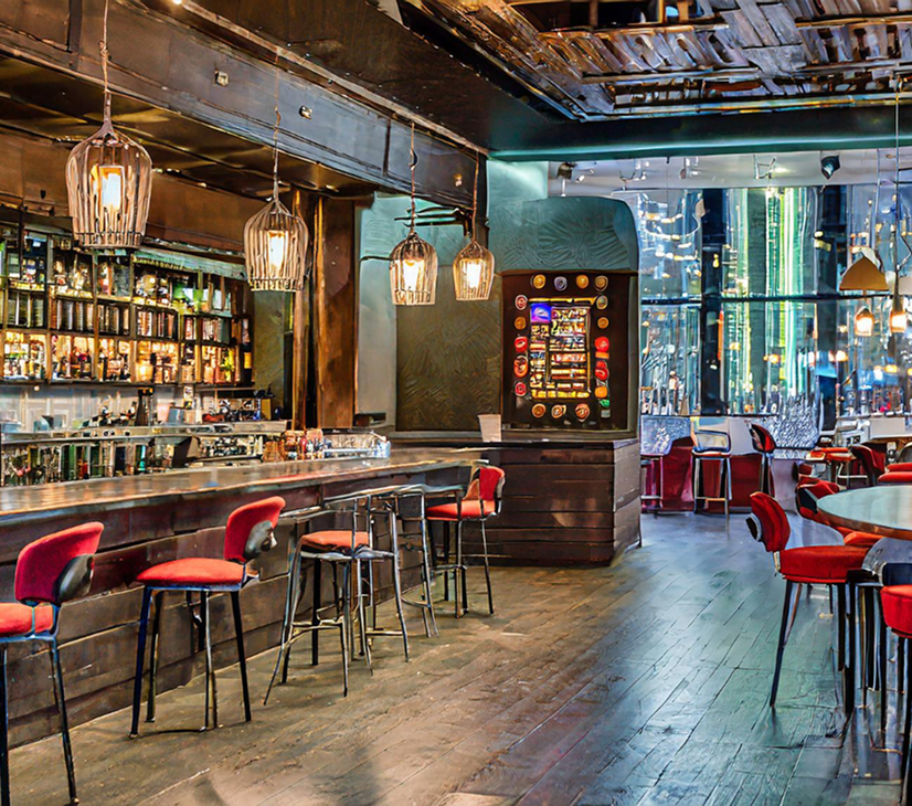 Interior of a stylish bar with red velvet chairs, wooden bar counter, and hanging pendant lights. A slot machine is visible in the background, and large windows reveal a cityscape with tall buildings.