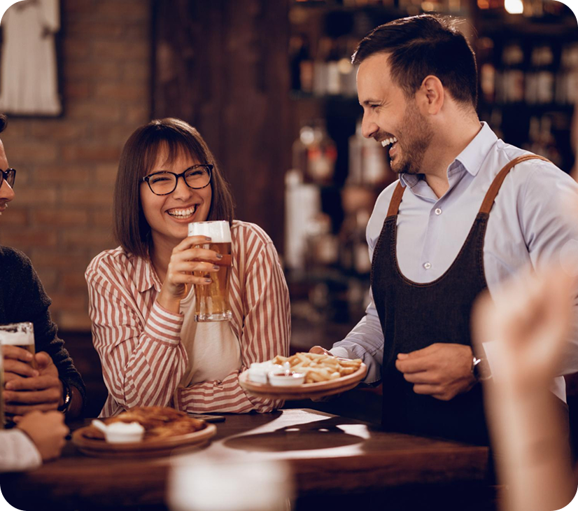 A bartender serving a plate of fries to a smiling woman wearing glasses in a cozy bar or pub setting.