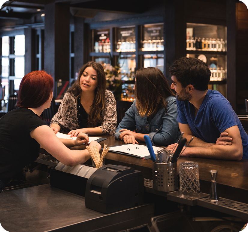A group of four young adults, two women and two men, sitting at a bar counter engaged in conversation with a bartender inside a dimly lit restaurant or bar with shelves of liquor bottles in the background.