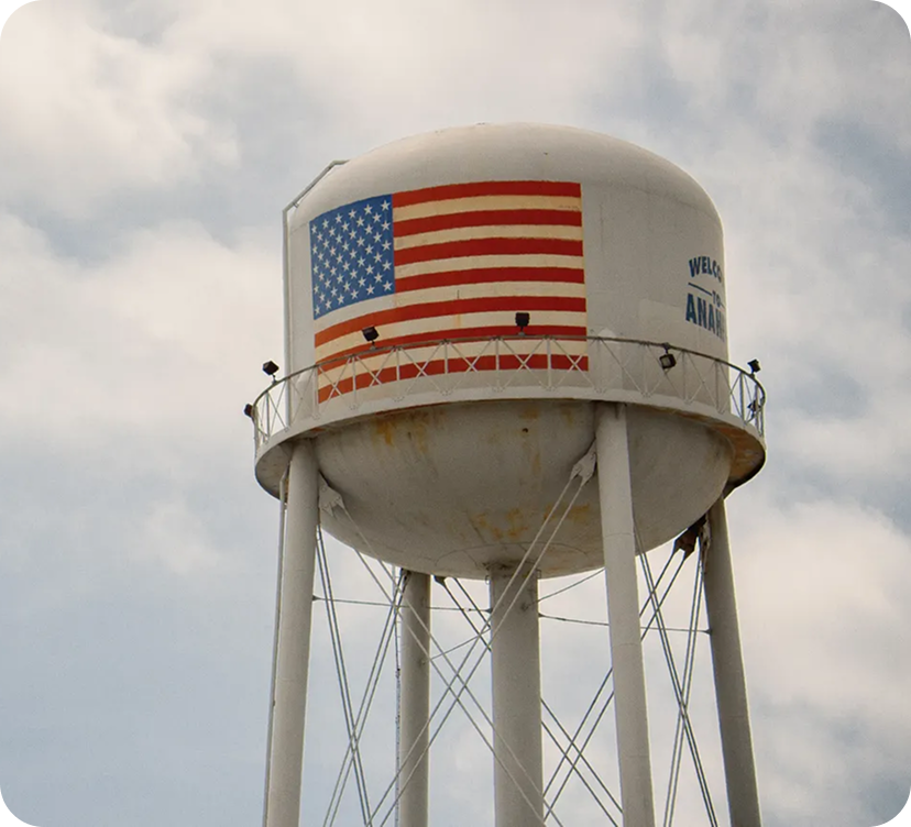 Water tower with an American flag and welcoming message, under cloudy sky.