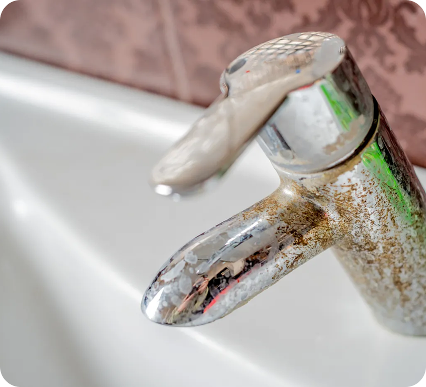 Close-up of a rusty, dirty bathroom faucet with water dripping from it, mounted above a white sink.