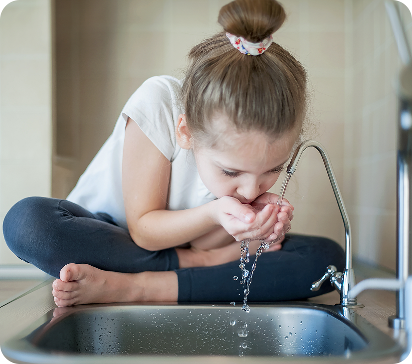 A young girl sitting cross-legged on a kitchen countertop, drinking water directly from a kitchen faucet with her hands cupped below.