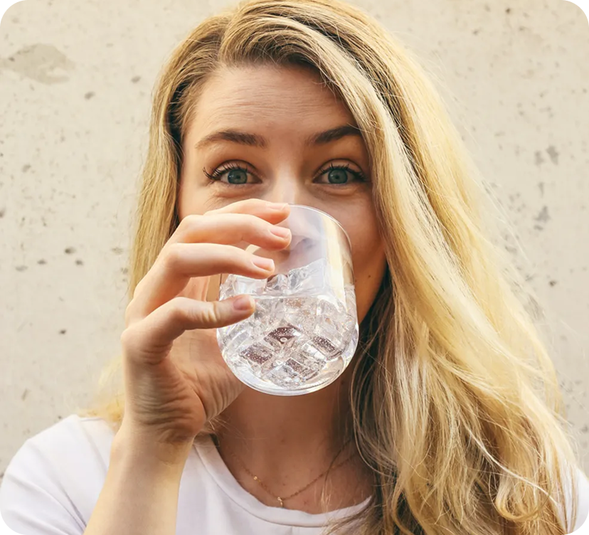 A woman with long blonde hair drinking from a glass of water, looking directly at the camera.