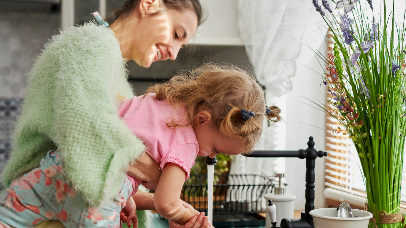 A woman and a young girl joyfully playing at a kitchen sink, with the woman helping the girl wash her hands.
