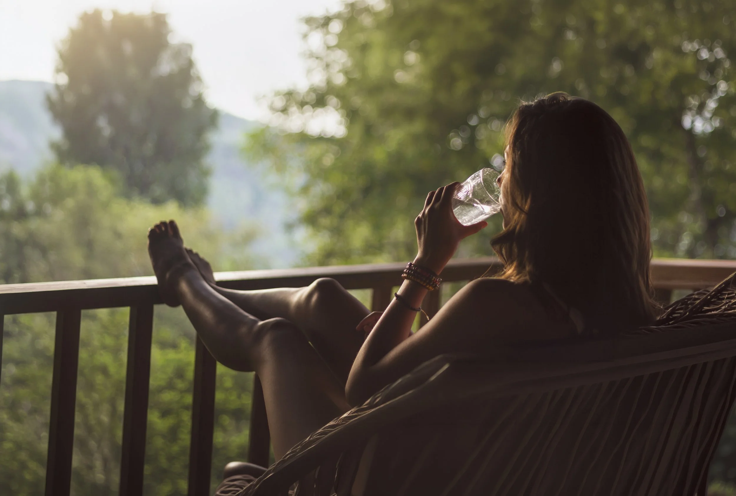 A woman relaxing on a porch, drinking from a clear glass, with lush green trees and sunlight in the background.