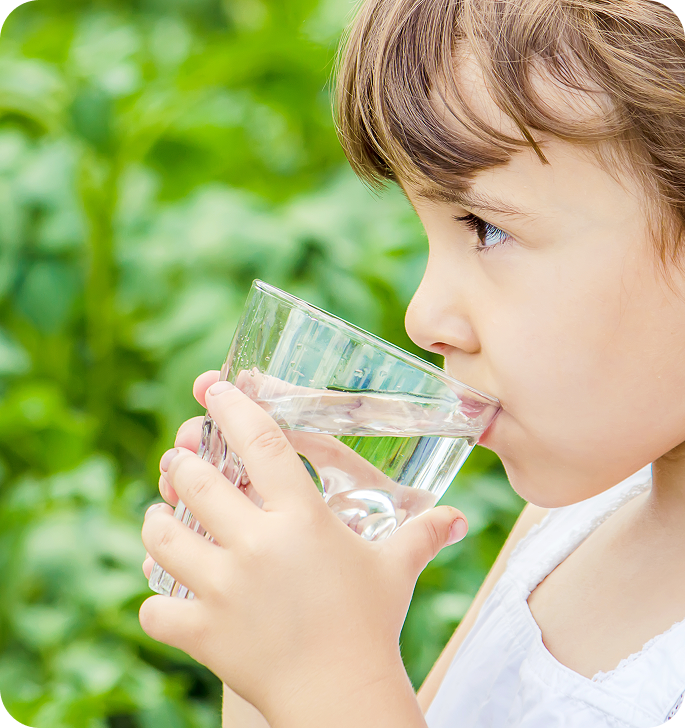 A young boy with light brown hair drinking from a clear glass of water outdoors, with green foliage in the background.