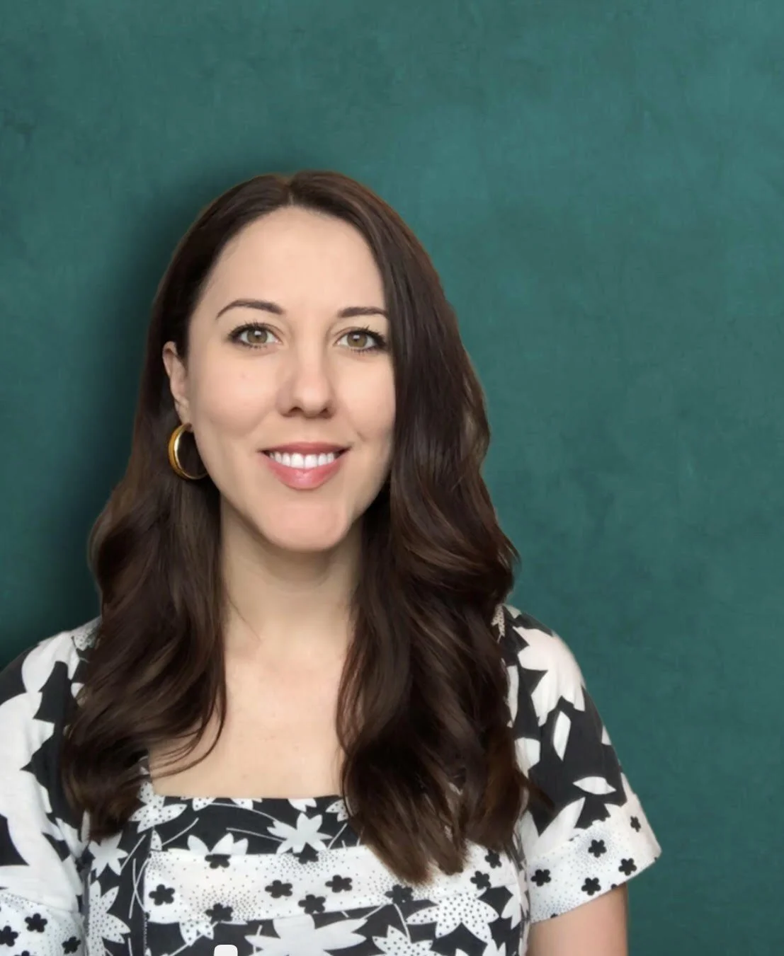 A smiling woman with long wavy brown hair, wearing a black and white patterned top and gold hoop earrings, standing against a teal background.