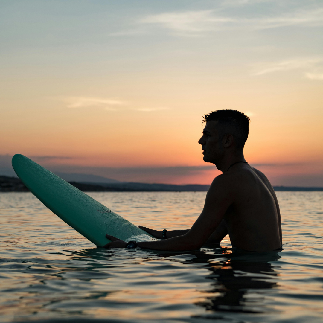 A man with short hair and no shirt holding a surfboard in the water during sunset.