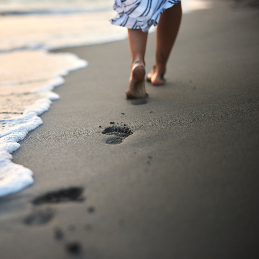 Person walking barefoot on the beach, leaving footprints in the sand near the shoreline with waves approaching.
