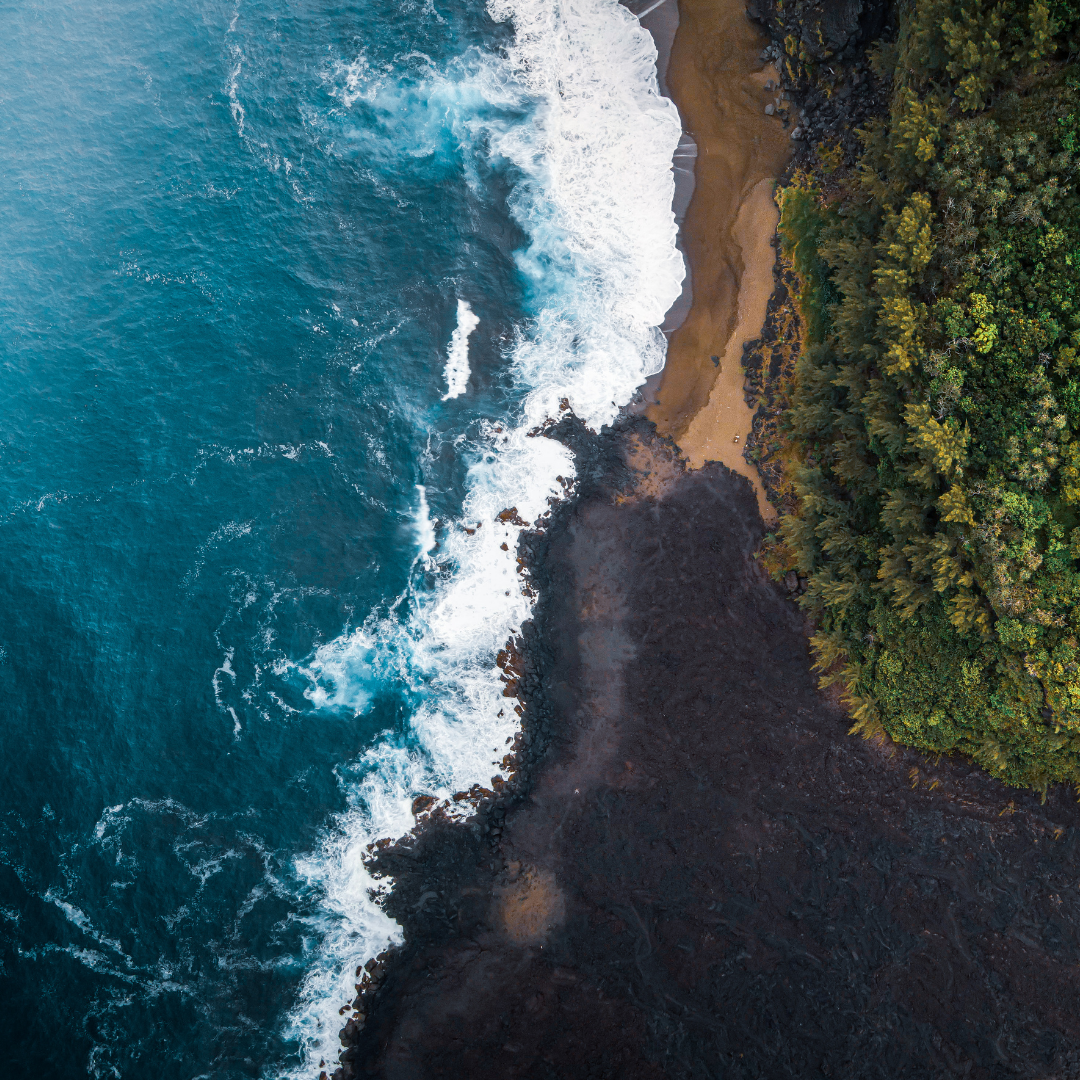 Aerial view of the coastline showing blue ocean waves crashing onto black volcanic rocks, sandy beach, and green forested area.