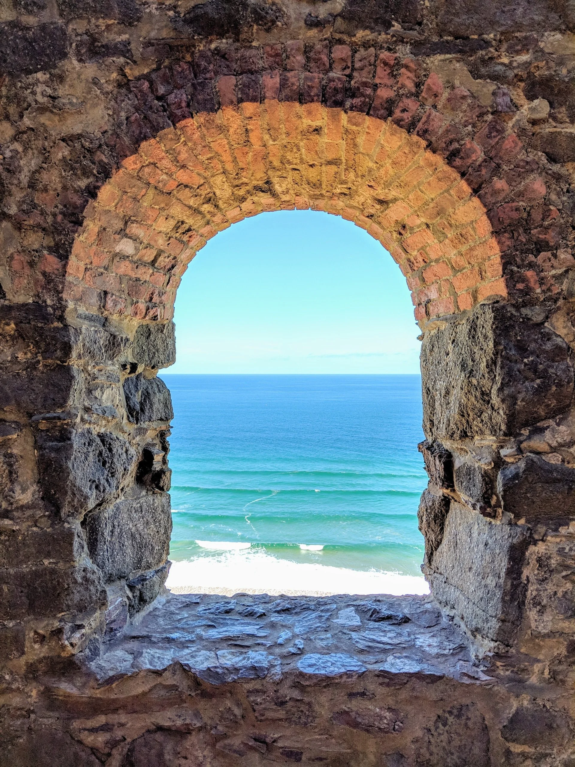 Looking through a stone and brick archway at the ocean with waves and a clear blue sky.