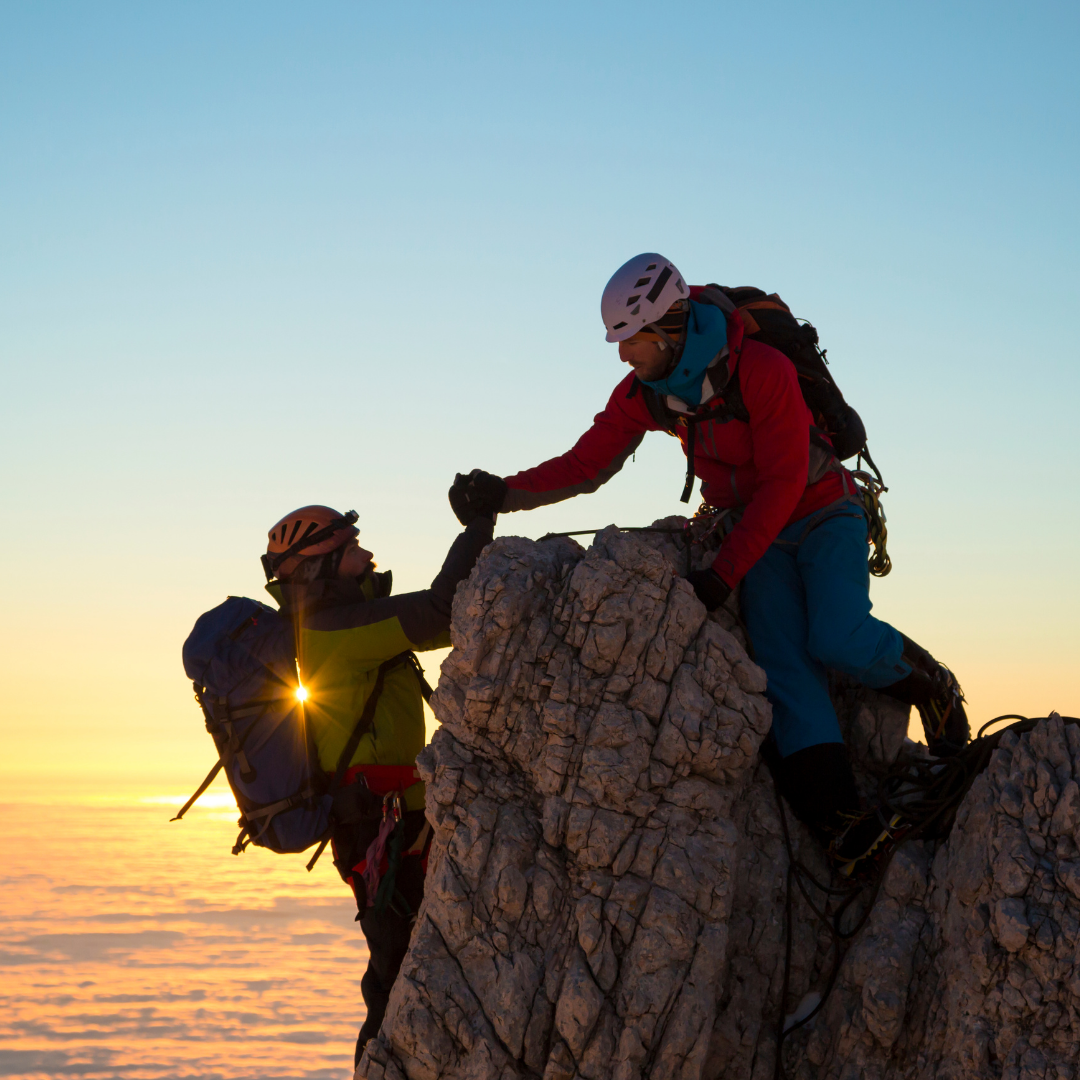 Two climbers helping each other on a rocky mountain during sunset, with clear sky and snow below.