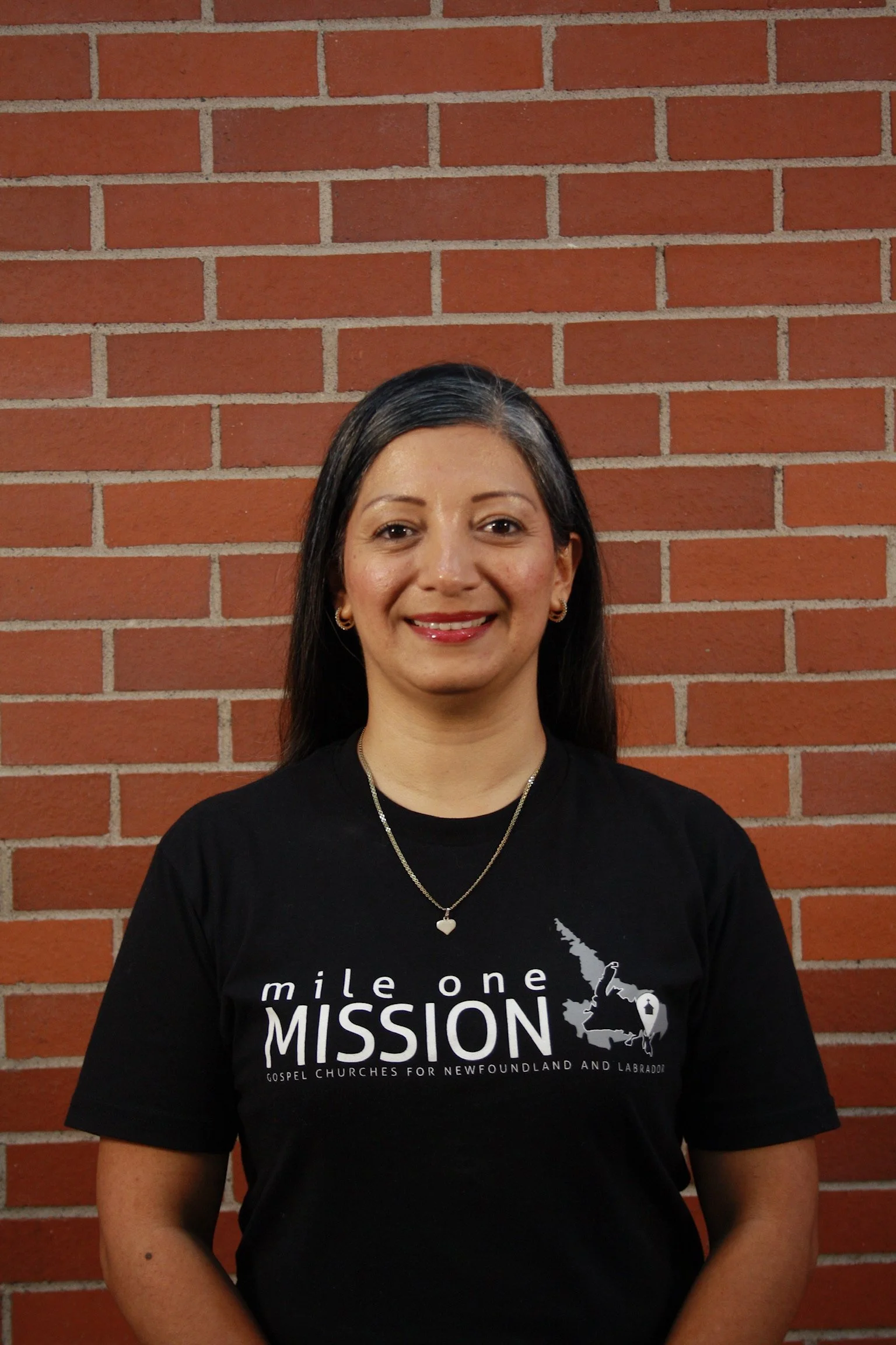 A woman with long dark hair, wearing earrings, smiling, standing in front of a red brick wall. She is wearing a black t-shirt with white text and a map of a region, and a necklace with a heart-shaped pendant.