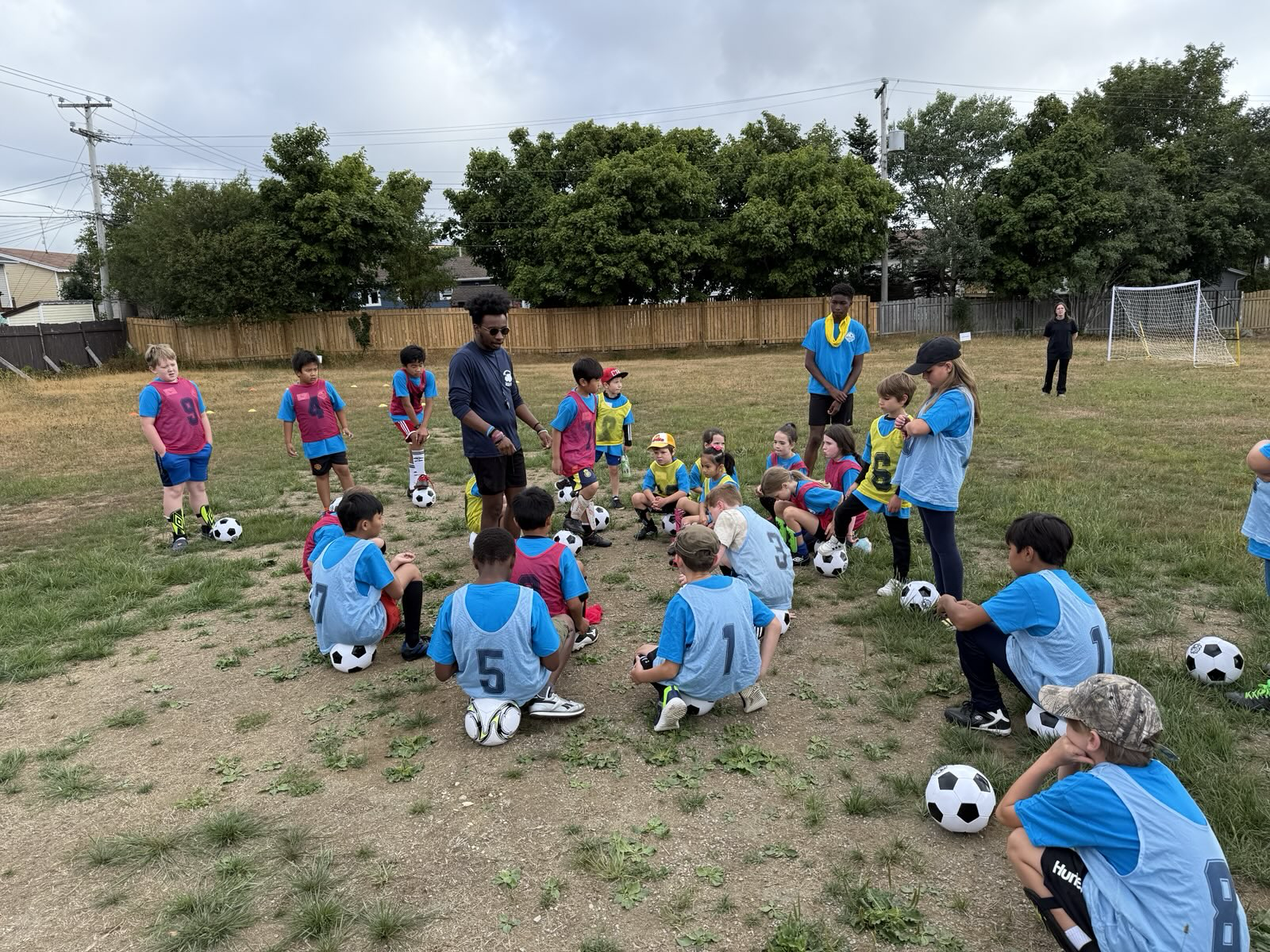 Group of children and coaches on a soccer field, with children sitting or standing with soccer balls, preparing for practice or a game.