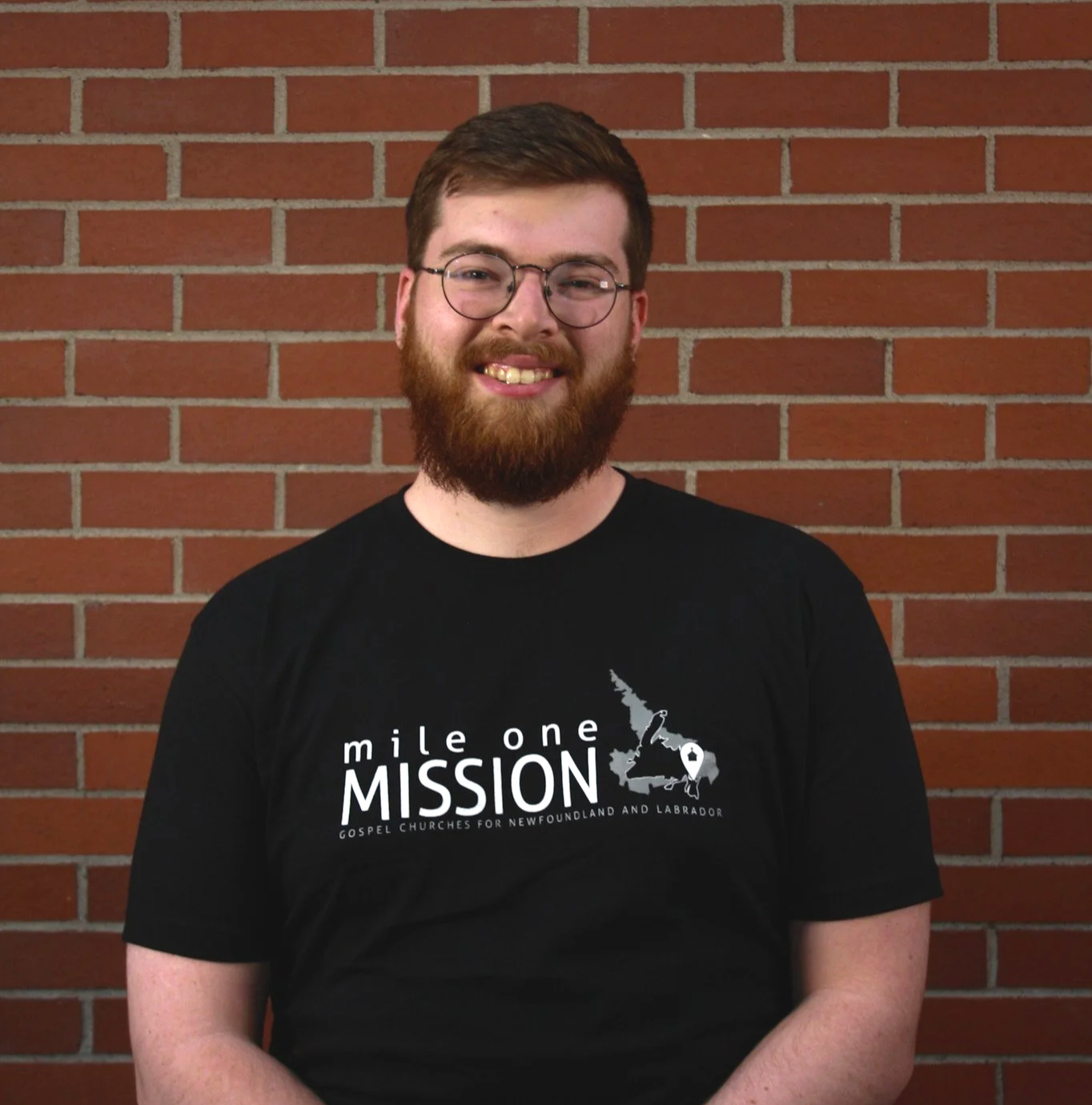 A young man with glasses and a beard, smiling, standing in front of a brick wall, wearing a black t-shirt with the text and graphics related to a mission for Newfoundland and Labrador.