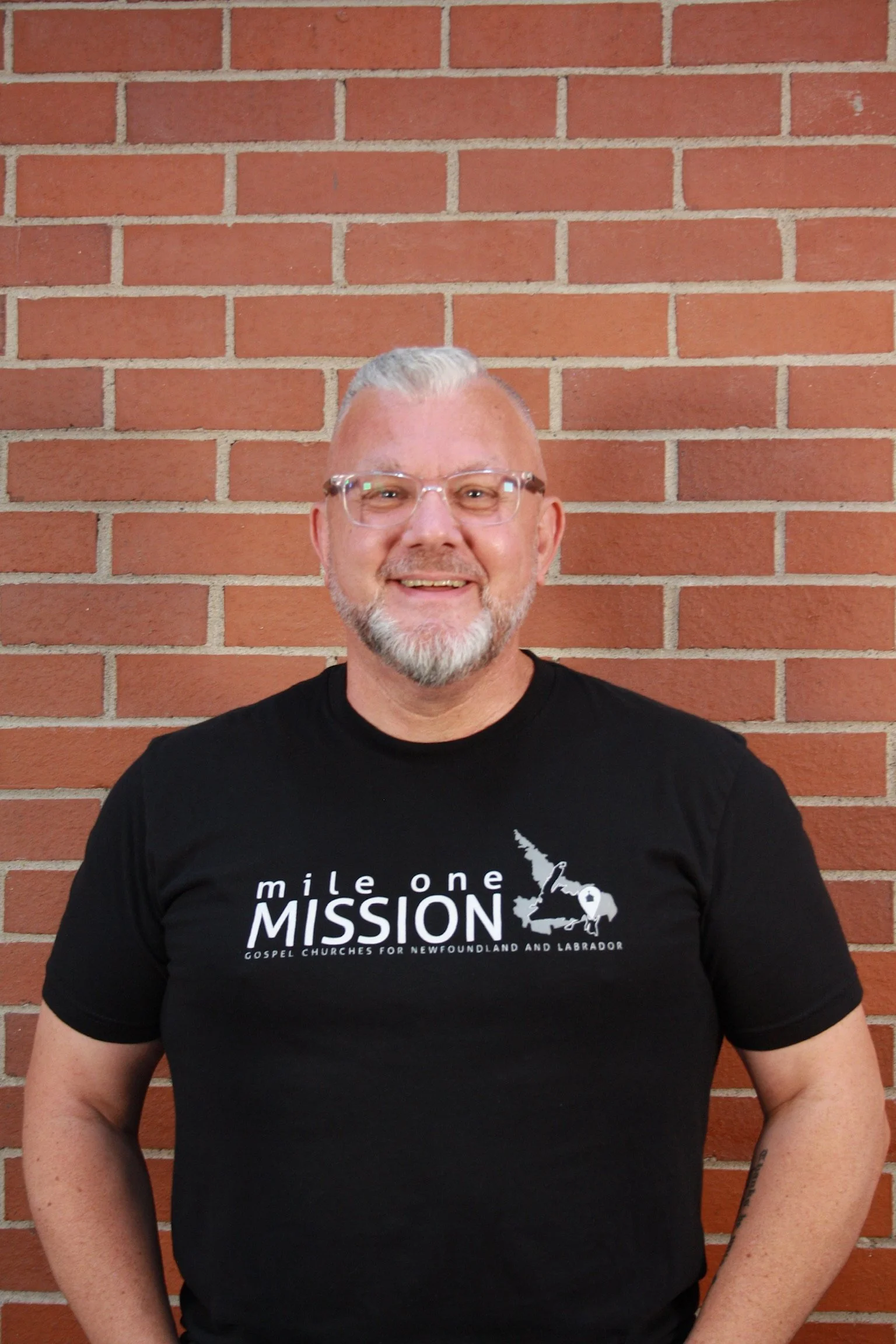 A smiling man with short gray hair and glasses standing in front of a red brick wall, wearing a black T-shirt with white text and graphics.