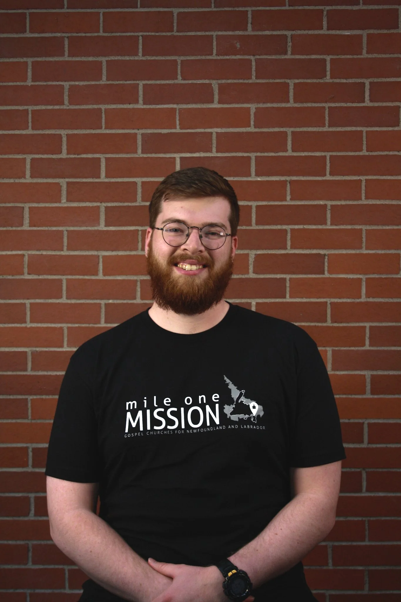 A man with glasses and a beard standing in front of a red brick wall, wearing a black t-shirt with a mission logo.