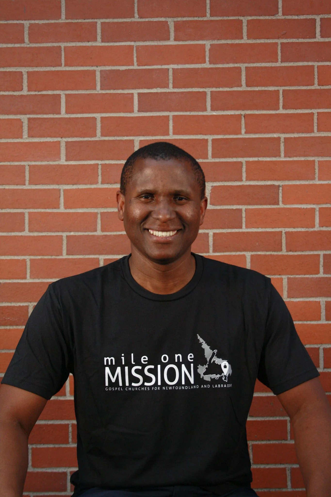 A smiling man with short hair, sitting in front of a red brick wall, wearing a black T-shirt with white text and graphics related to a mission or church.