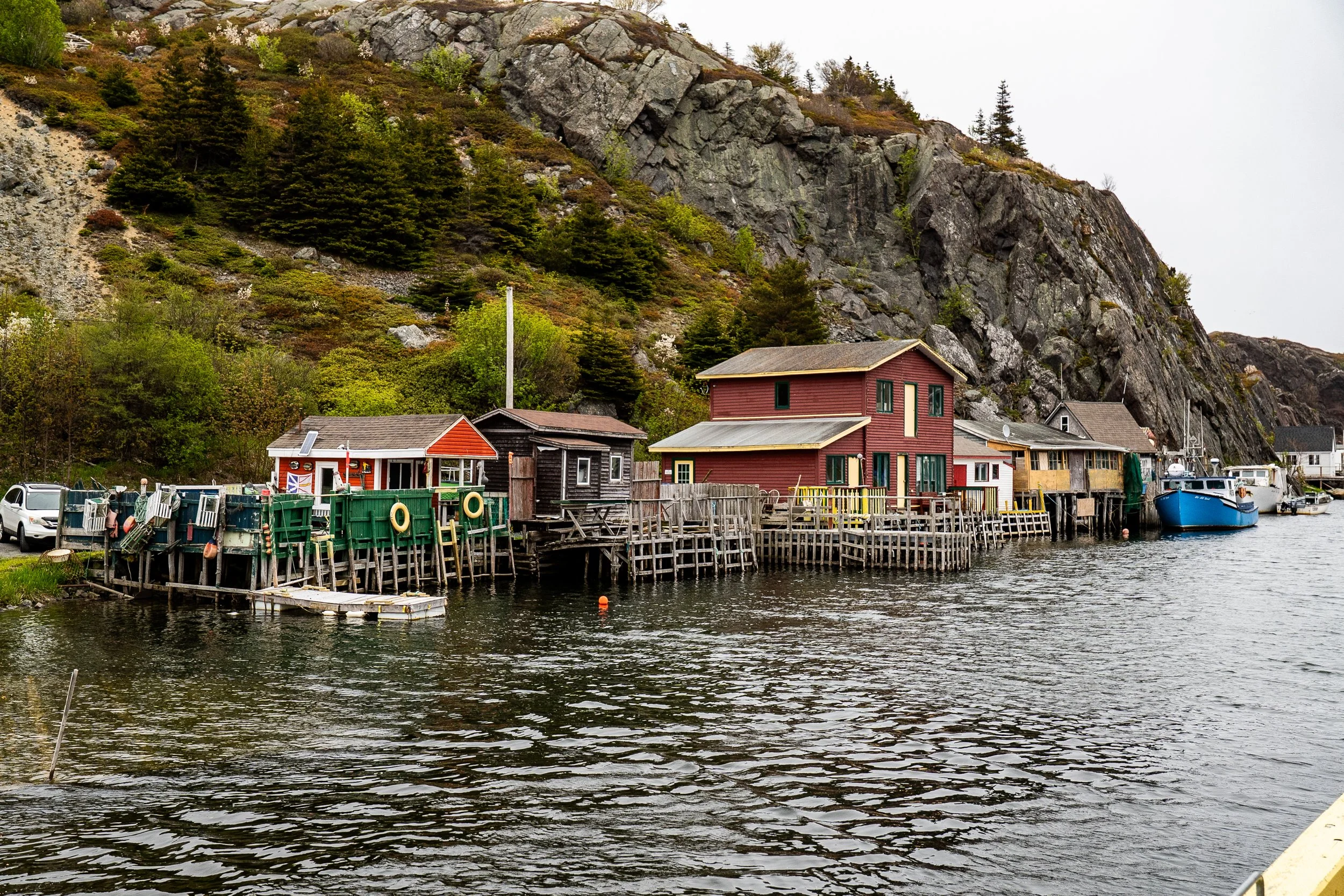 A small harbor with colorful houses along the waterfront, surrounded by rocky, forested mountains under an overcast sky.