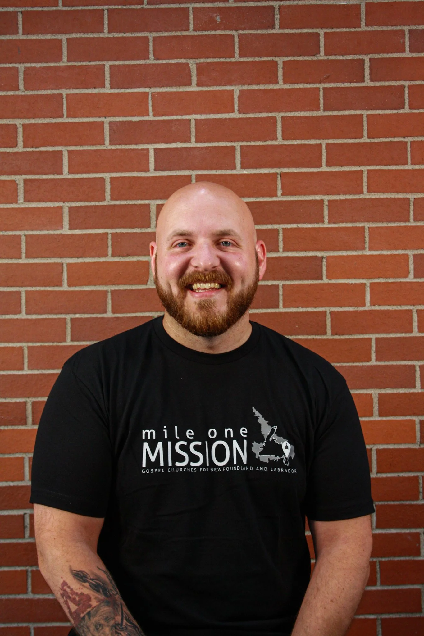 A smiling man with a bald head, full beard, and tattooed arm, wearing a black t-shirt with a map of Newfoundland and Labrador and the words "mile one MISSION gospel churches for Newfoundland and Labrador," standing in front of a red brick wall.