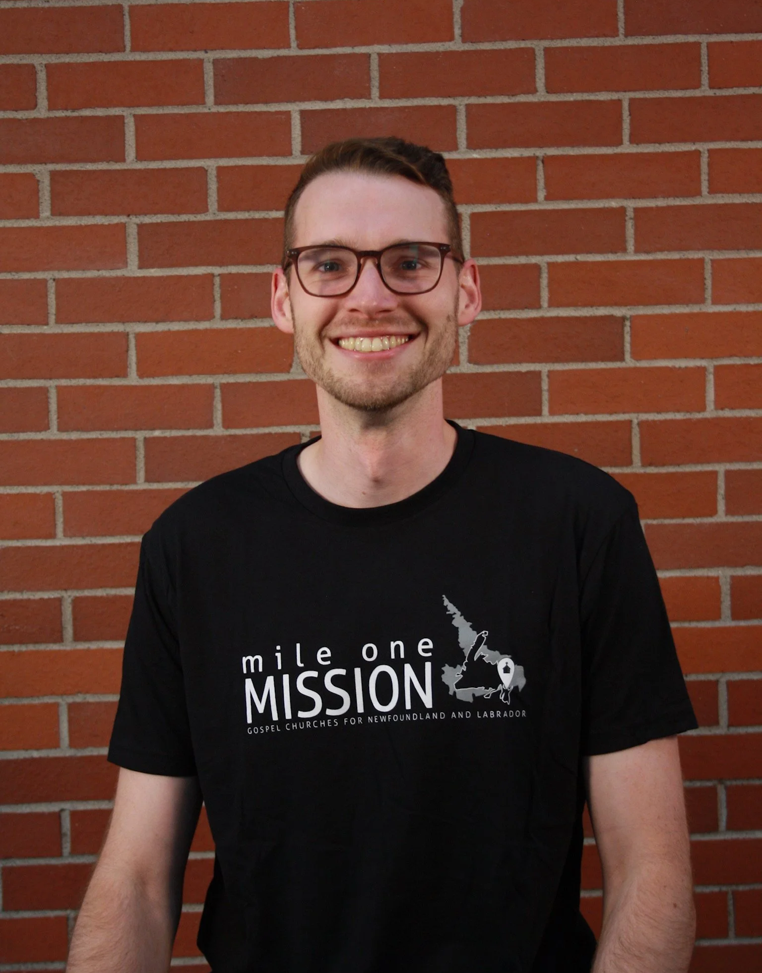A young man with short brown hair, glasses, and a beard, smiling, standing in front of a red brick wall, wearing a black T-shirt that says 'mile one MISSION' and includes an outline of Newfoundland and Labrador.