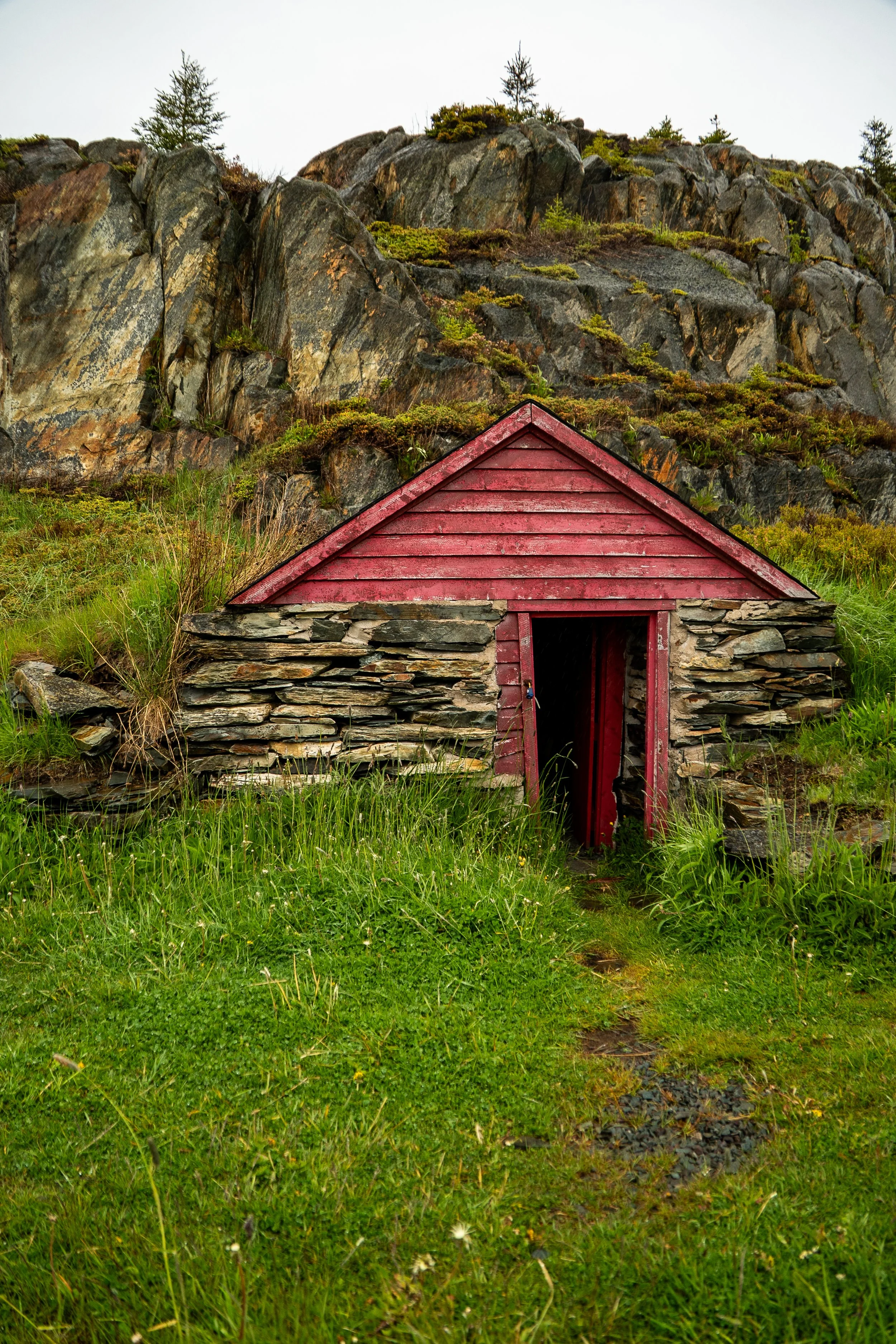 A small stone cottage with a red wooden roof and an open doorway, surrounded by green grass and located at the base of a rocky, moss-covered mountain.