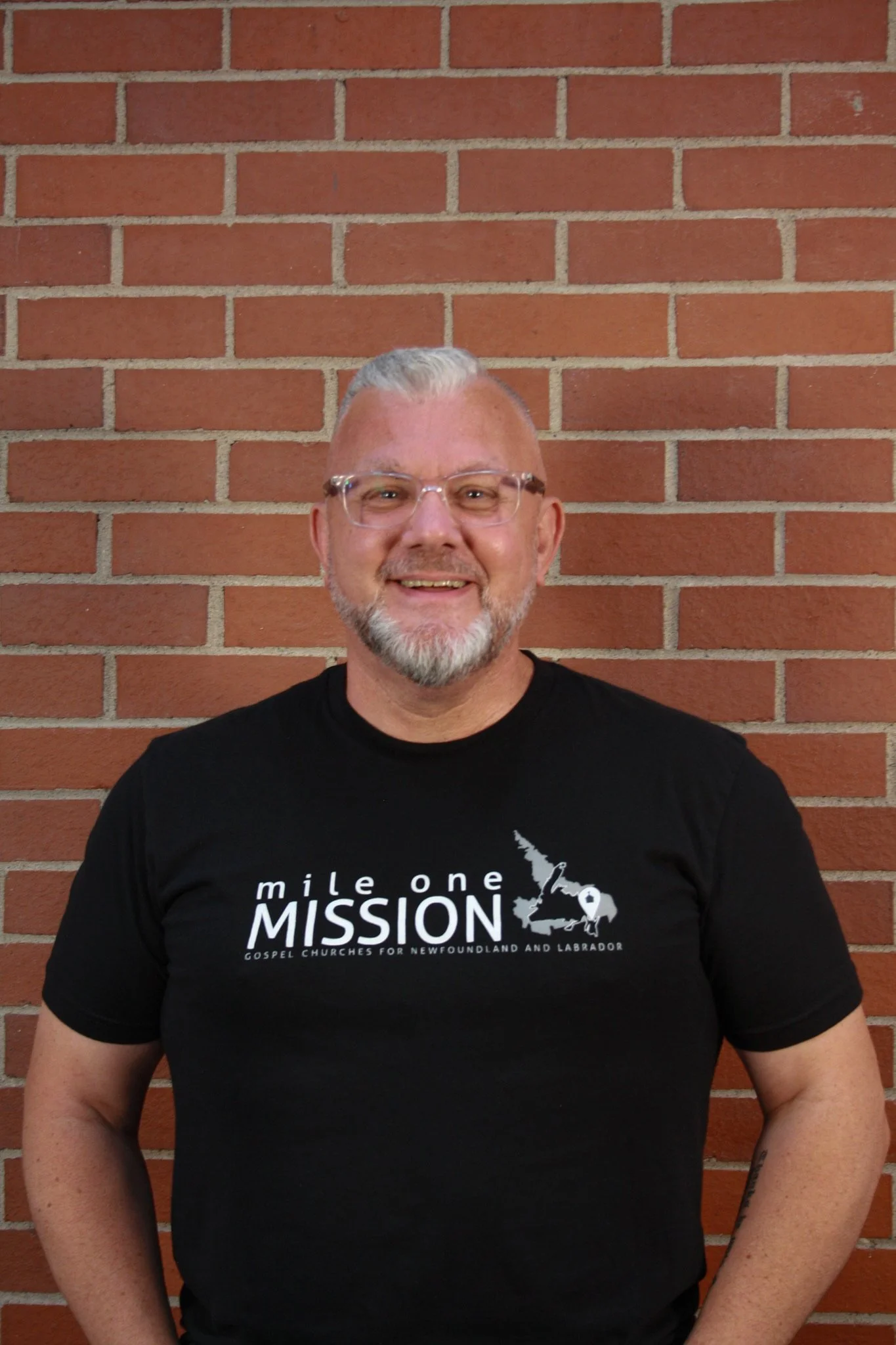 A smiling man with glasses and a short gray beard stands in front of a red brick wall. He is wearing a black t-shirt with white text about a gospel church mission in Newfoundland and Labrador.