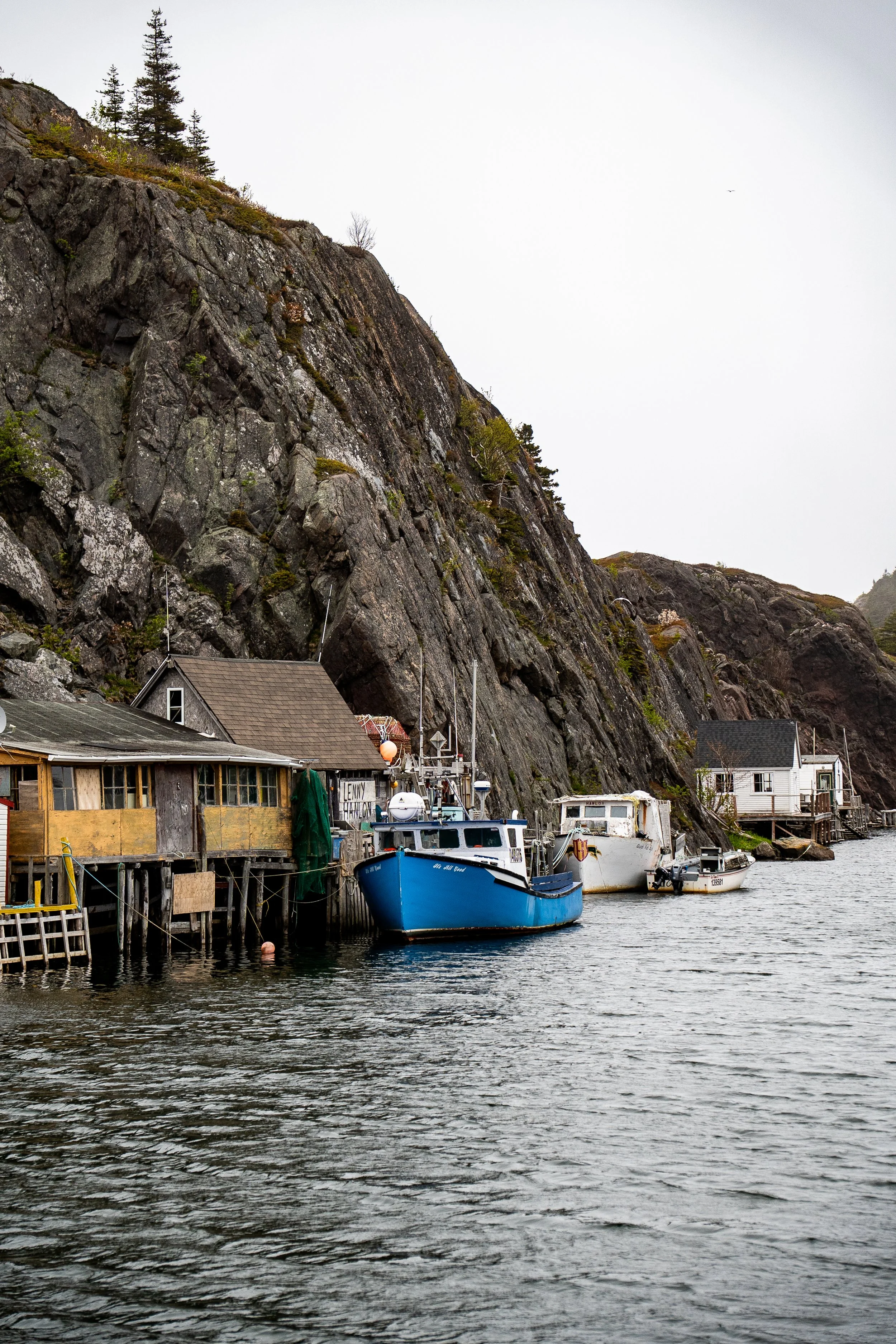 A harbor with several boats docked along wooden piers in front of a rocky hillside with sparse trees, under an overcast sky.