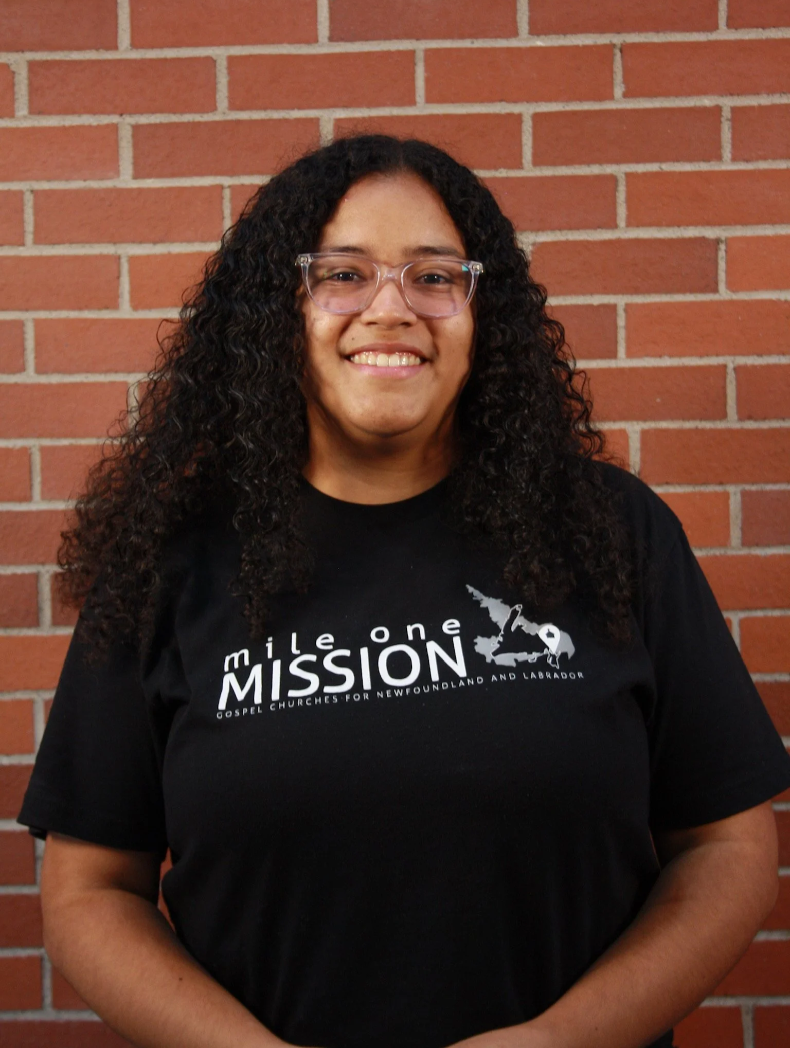 A woman with curly dark hair wearing glasses and a black T-shirt standing in front of a brick wall.