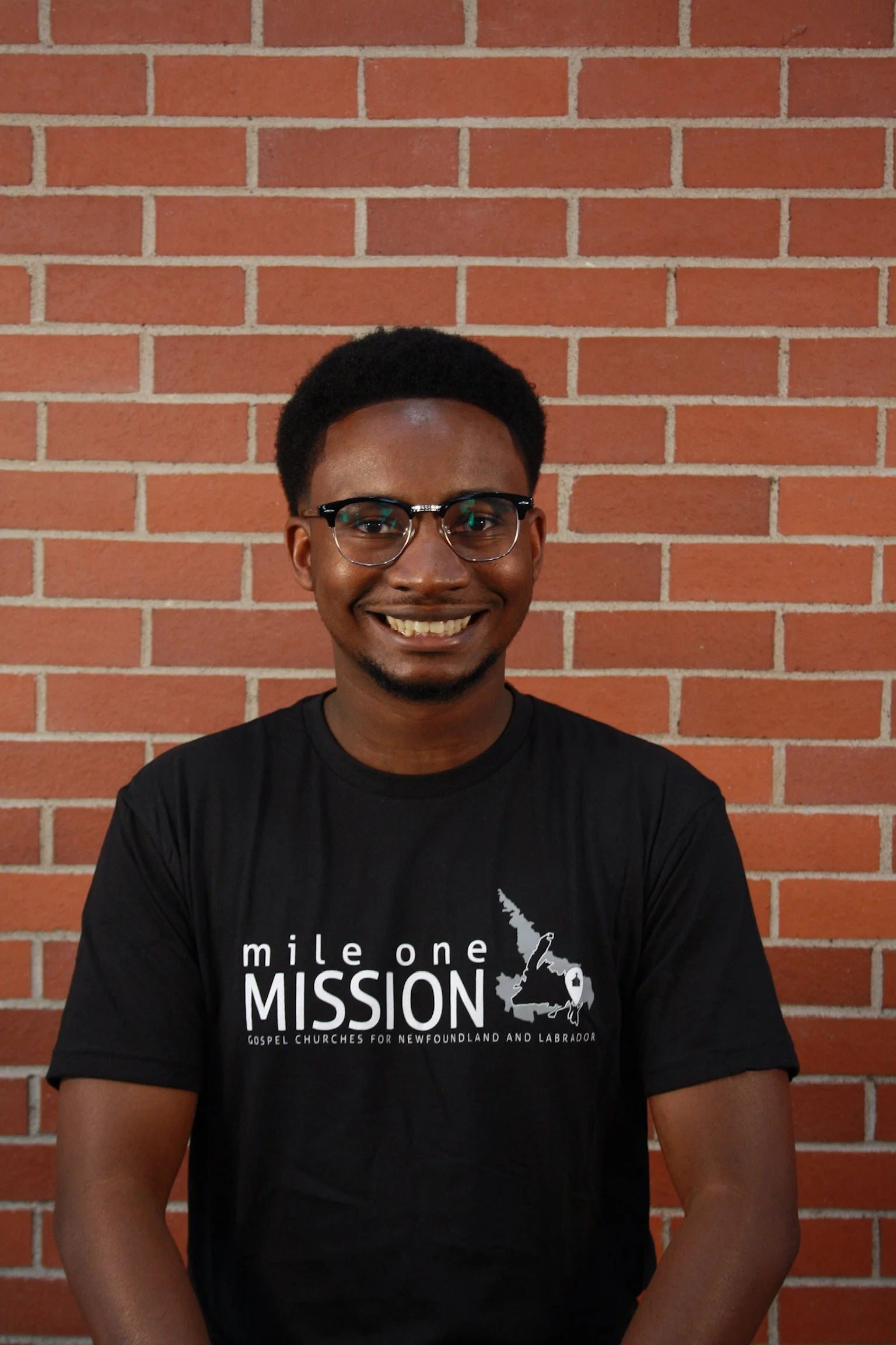A young man with glasses smiling in front of a brick wall, wearing a black T-shirt that reads 'mile one MISSION Gospel Churches for Newfoundland and Labrador' with a map of Newfoundland and Labrador.