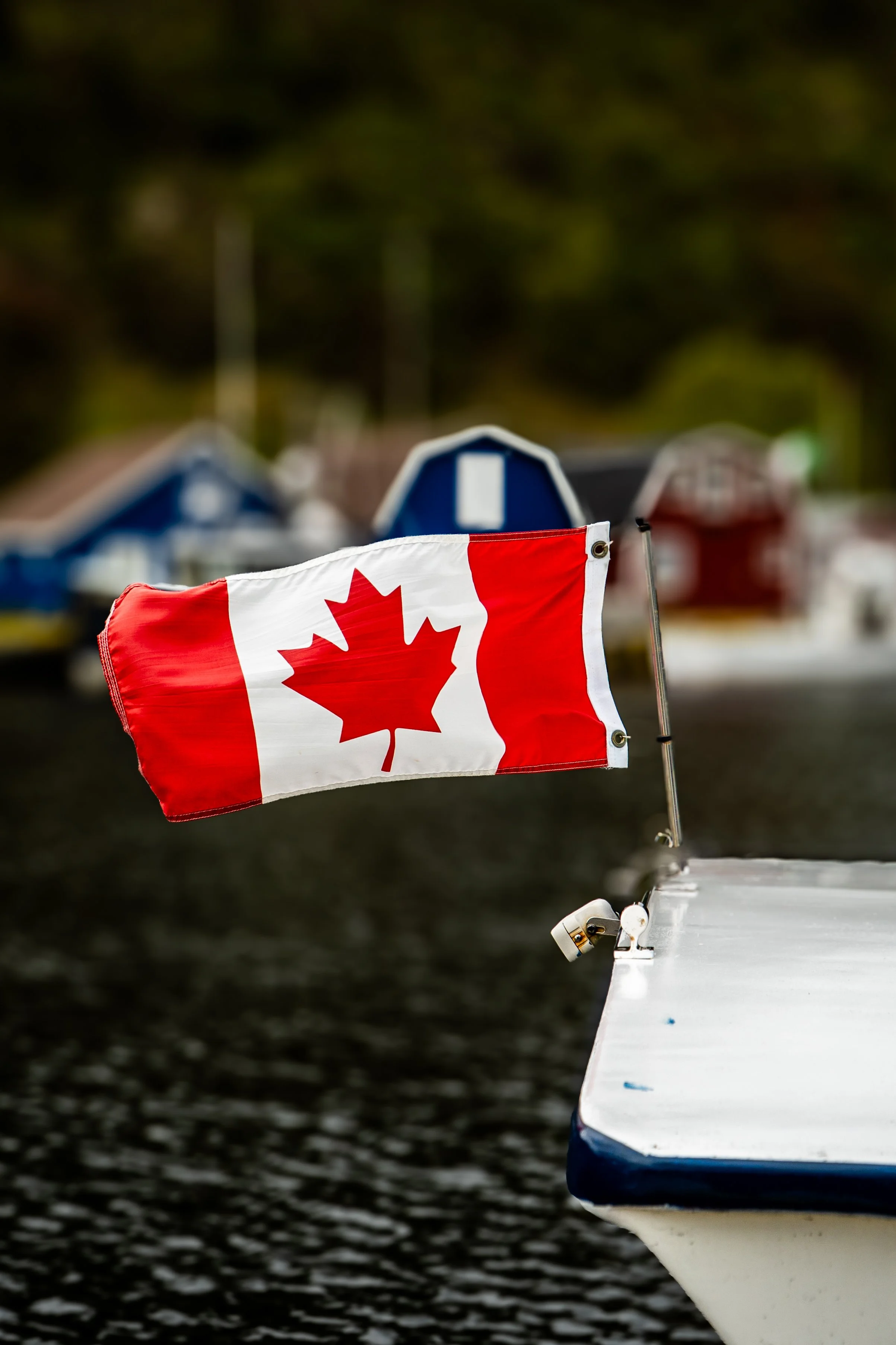 Close-up of a Canadian flag attached to the bow of a boat, with water in the background and blurred houses or buildings.