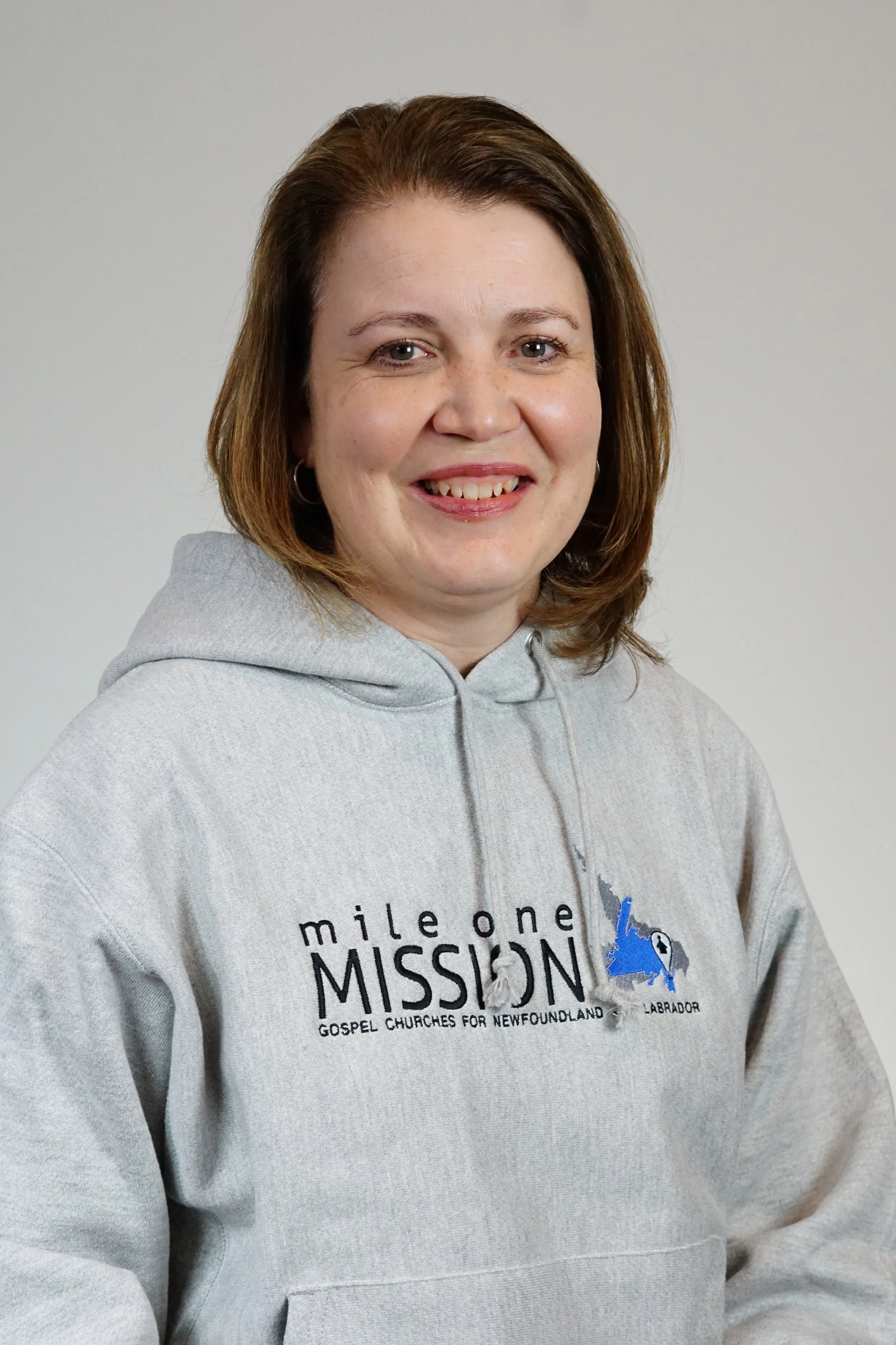 A woman with shoulder-length brown hair wearing a light gray hoodie with a logo for Mile One Mission, a gospel church in Newfoundland and Labrador, standing against a plain, light-colored background.