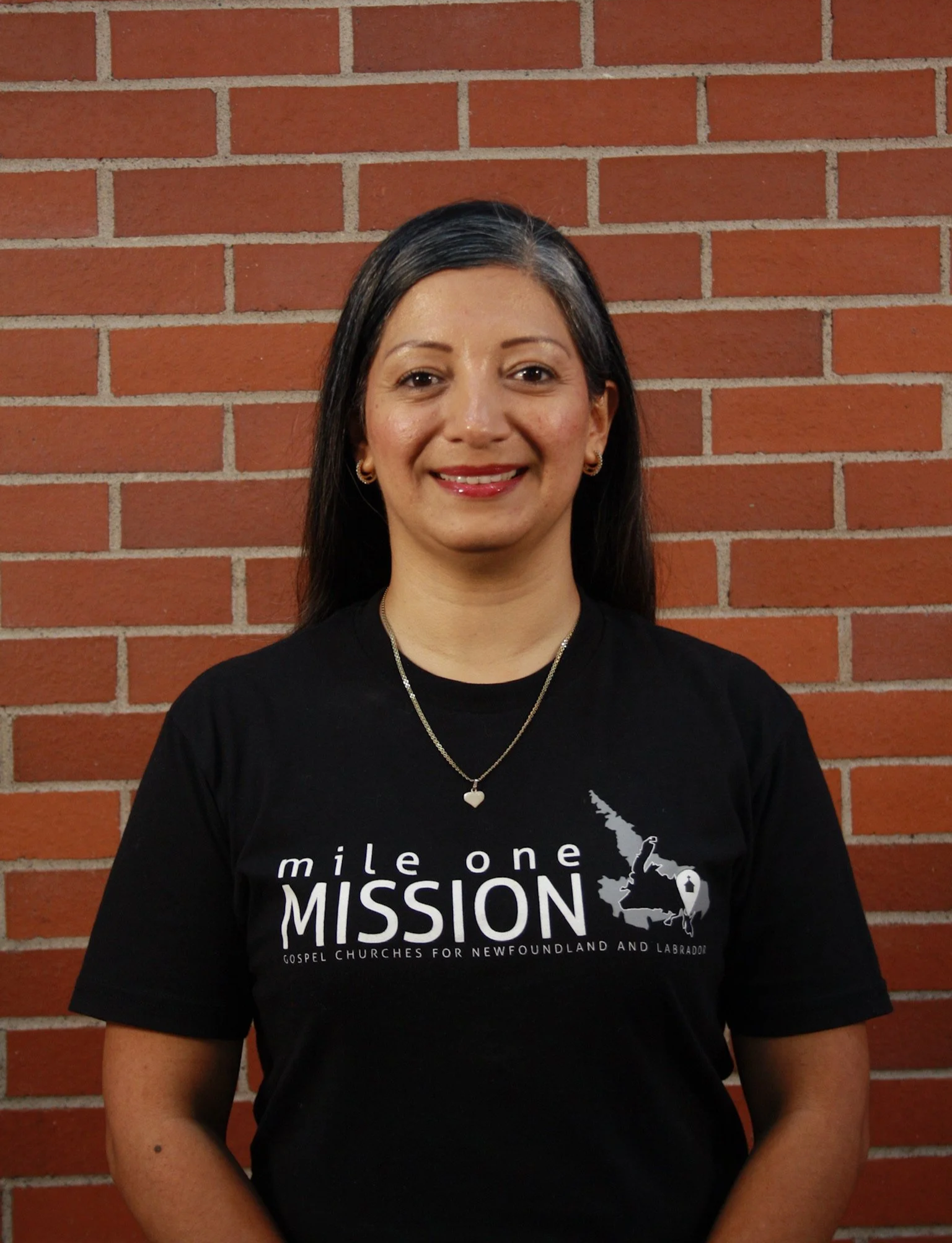 A woman with dark hair and earrings smiling in front of a brick wall. She wears a black T-shirt with white text about a mission and gospel churches in Newfoundland and Labrador.