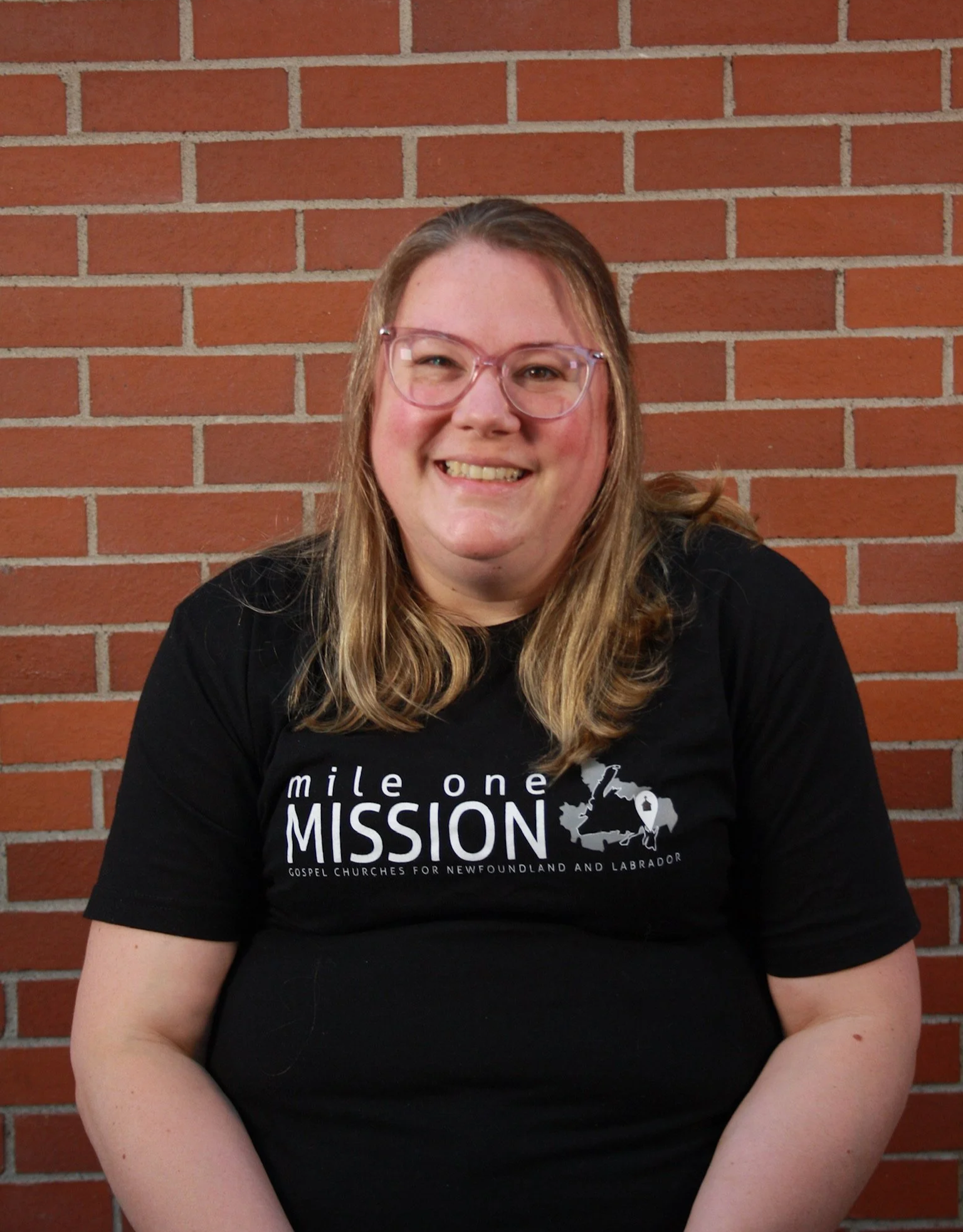 A woman with long light brown hair wearing glasses and a black t-shirt, smiling while sitting in front of a red brick wall.