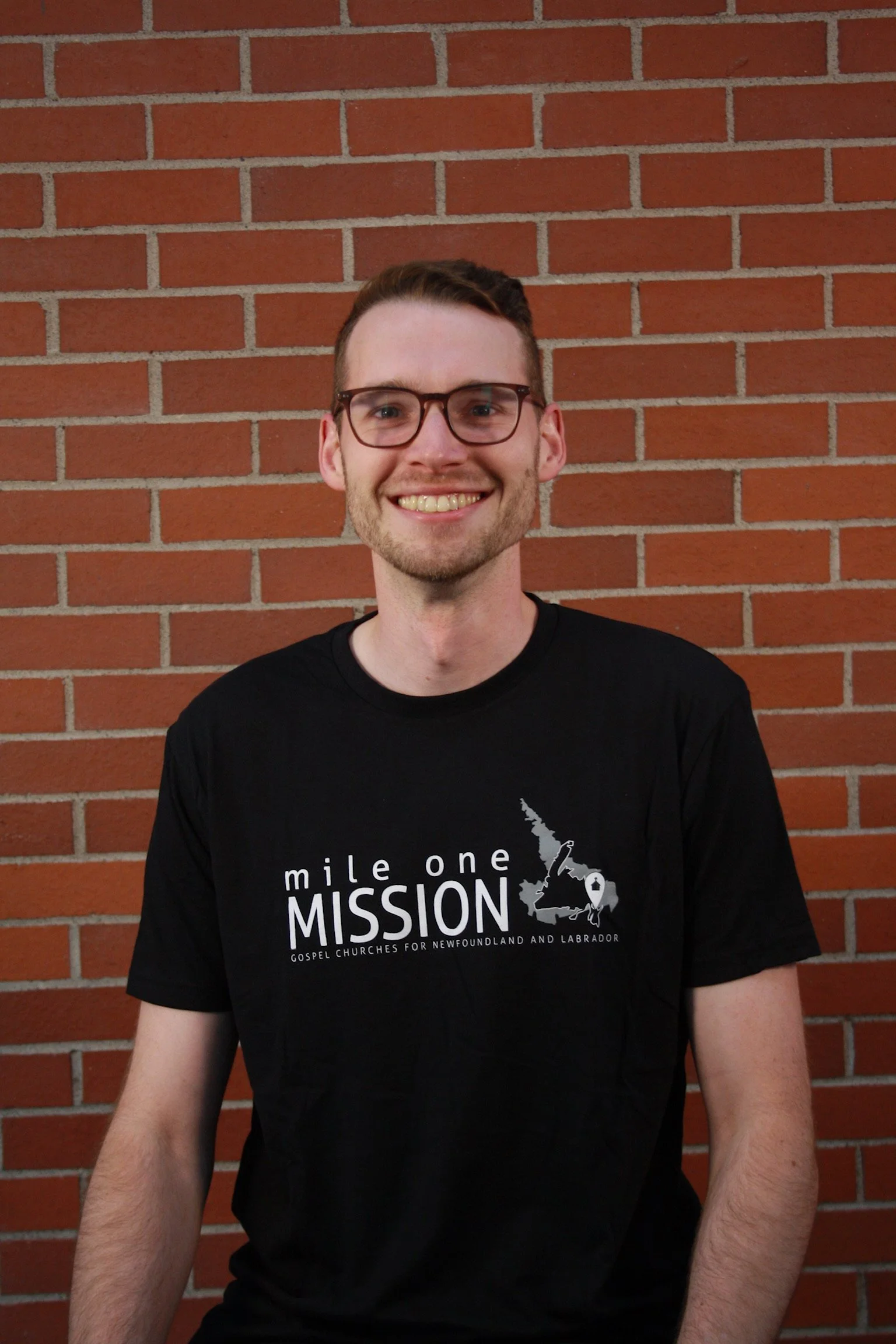 A smiling young man with glasses standing in front of a brick wall, wearing a black t-shirt with white text and a graphic related to a church mission in Newfoundland and Labrador.