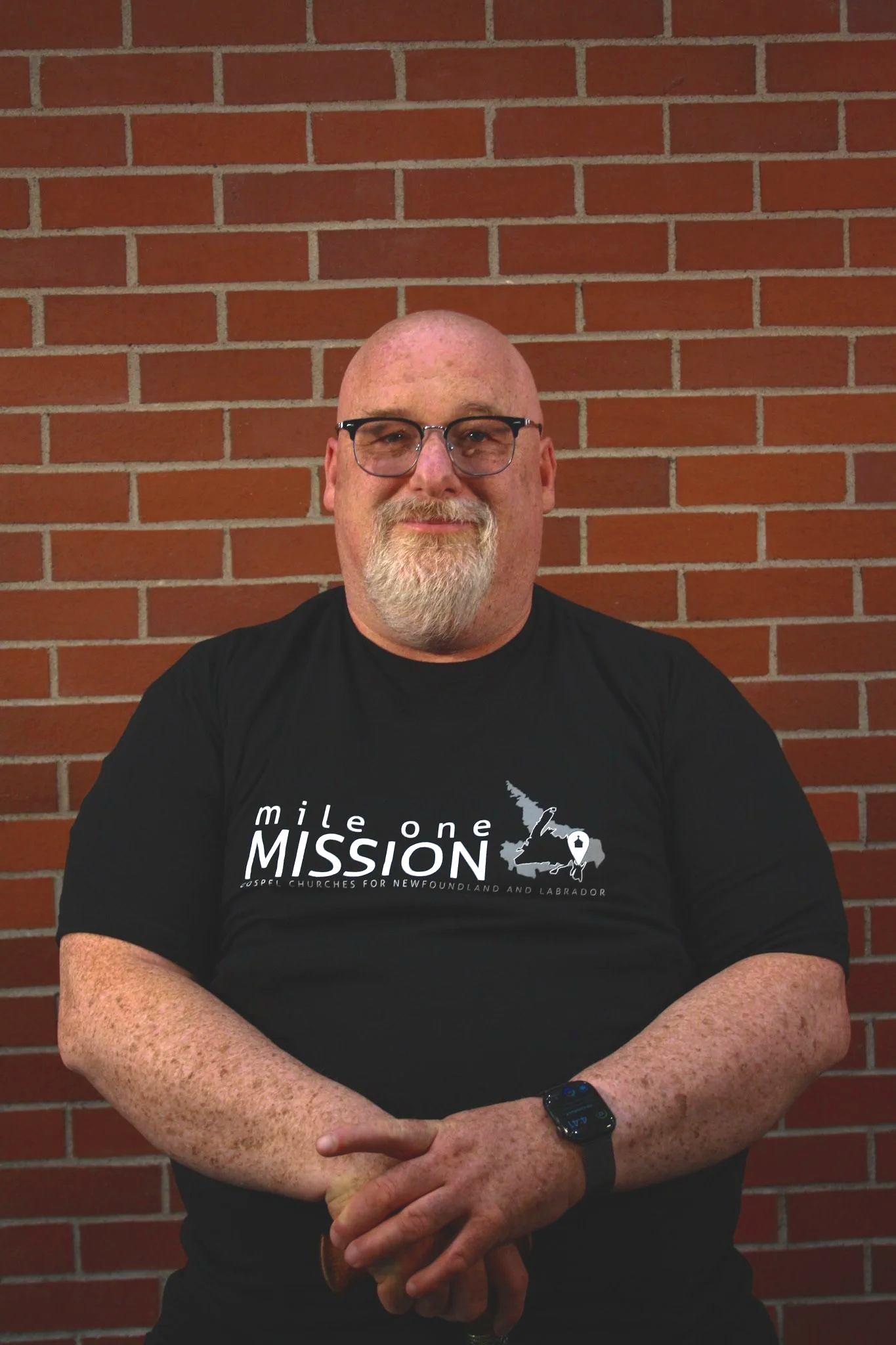 A middle-aged man with a shaved head, glasses, and a beard, wearing a black t-shirt with the words 'mile one MISSION' and an outline of Newfoundland and Labrador, standing in front of a red brick wall.