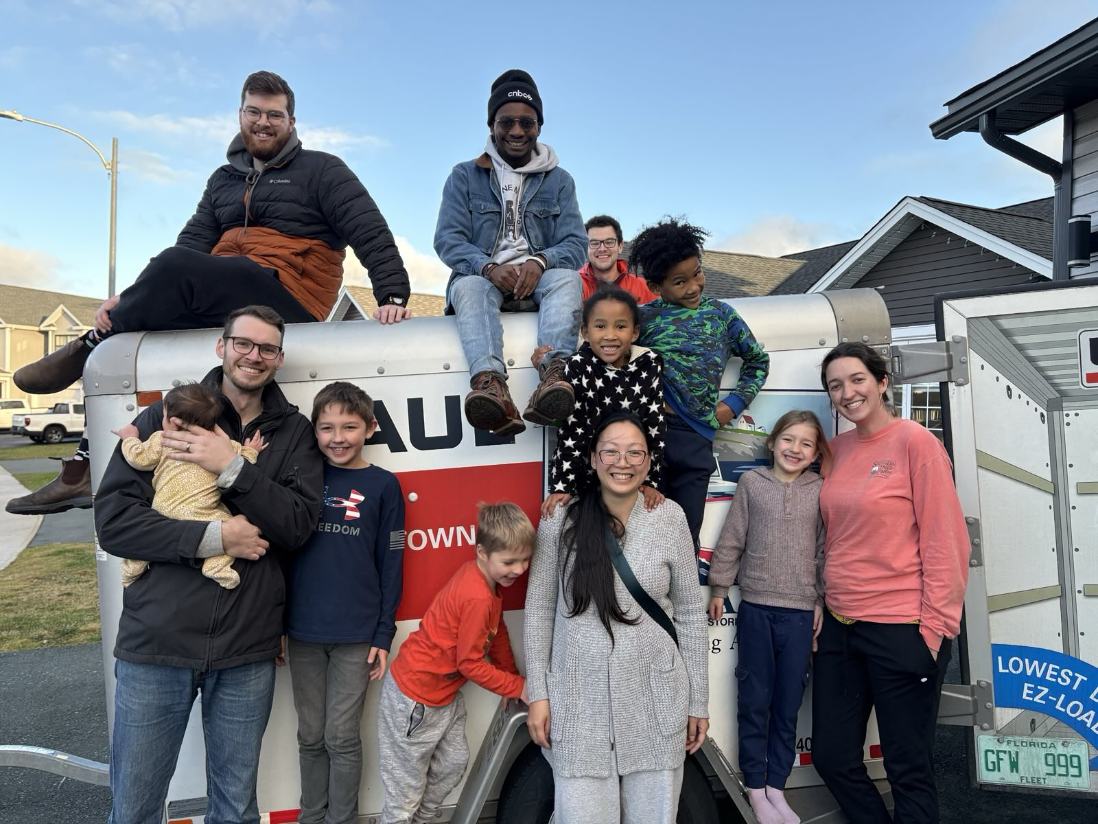 Group of people, including children and adults, gathered around a U-Haul truck, smiling and posing for a photo outdoors in a neighborhood with houses in the background.