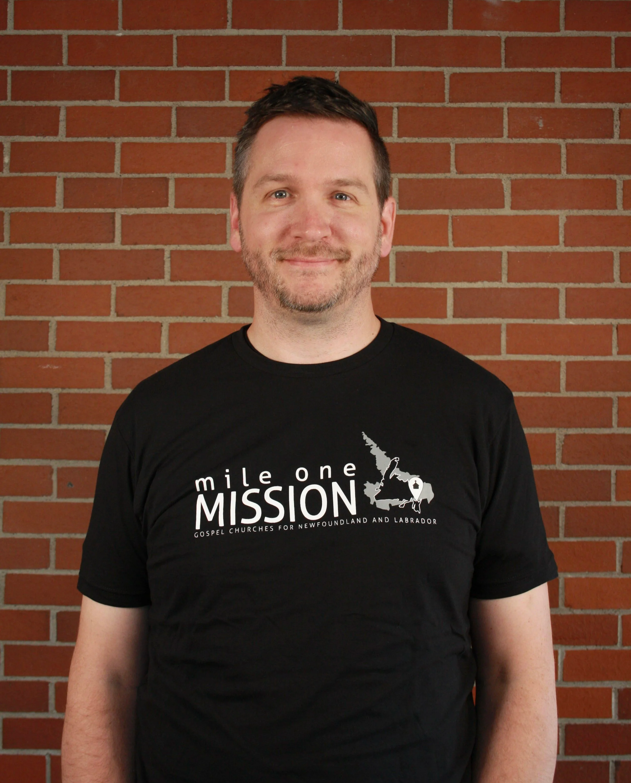 A man with short brown hair and light facial hair smiling and wearing a black t-shirt with white text and a graphic of Canada, standing in front of a brick wall.