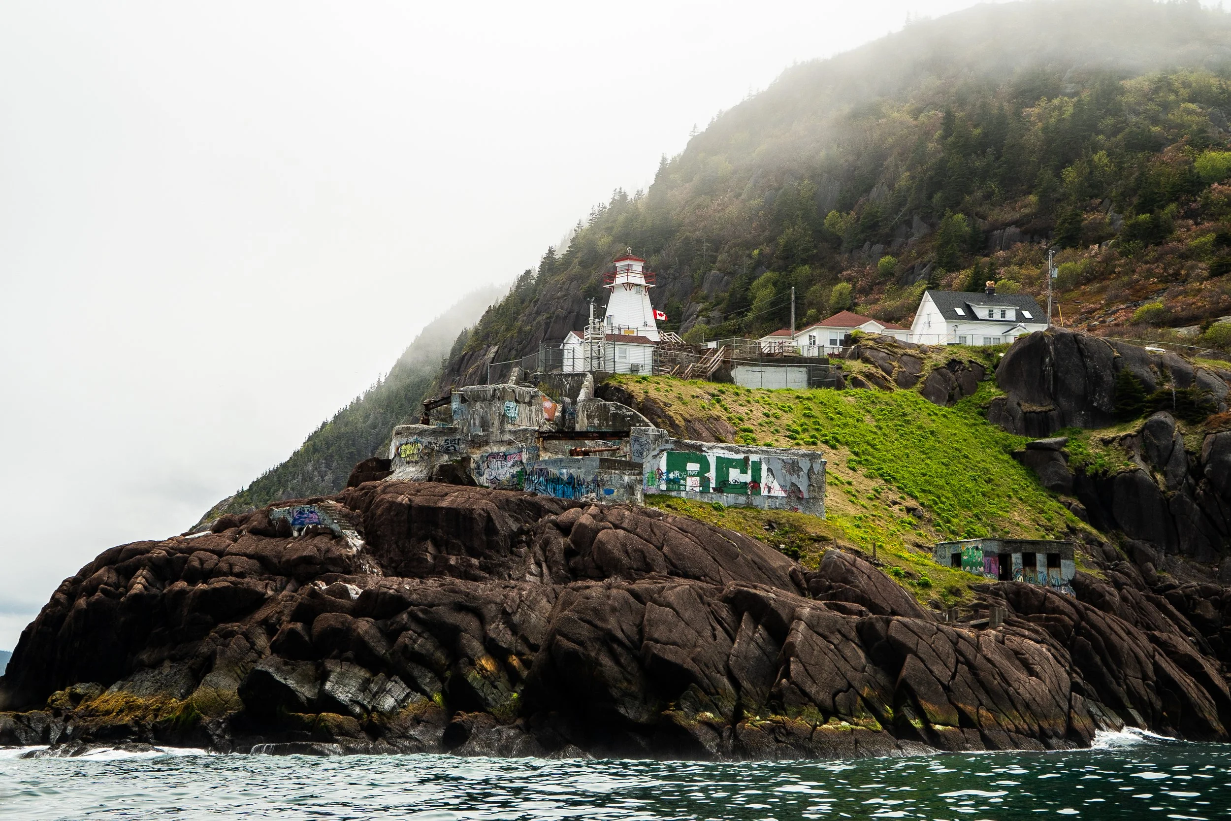A lighthouse on a rocky shoreline with buildings nearby, surrounded by lush green hills and fog.