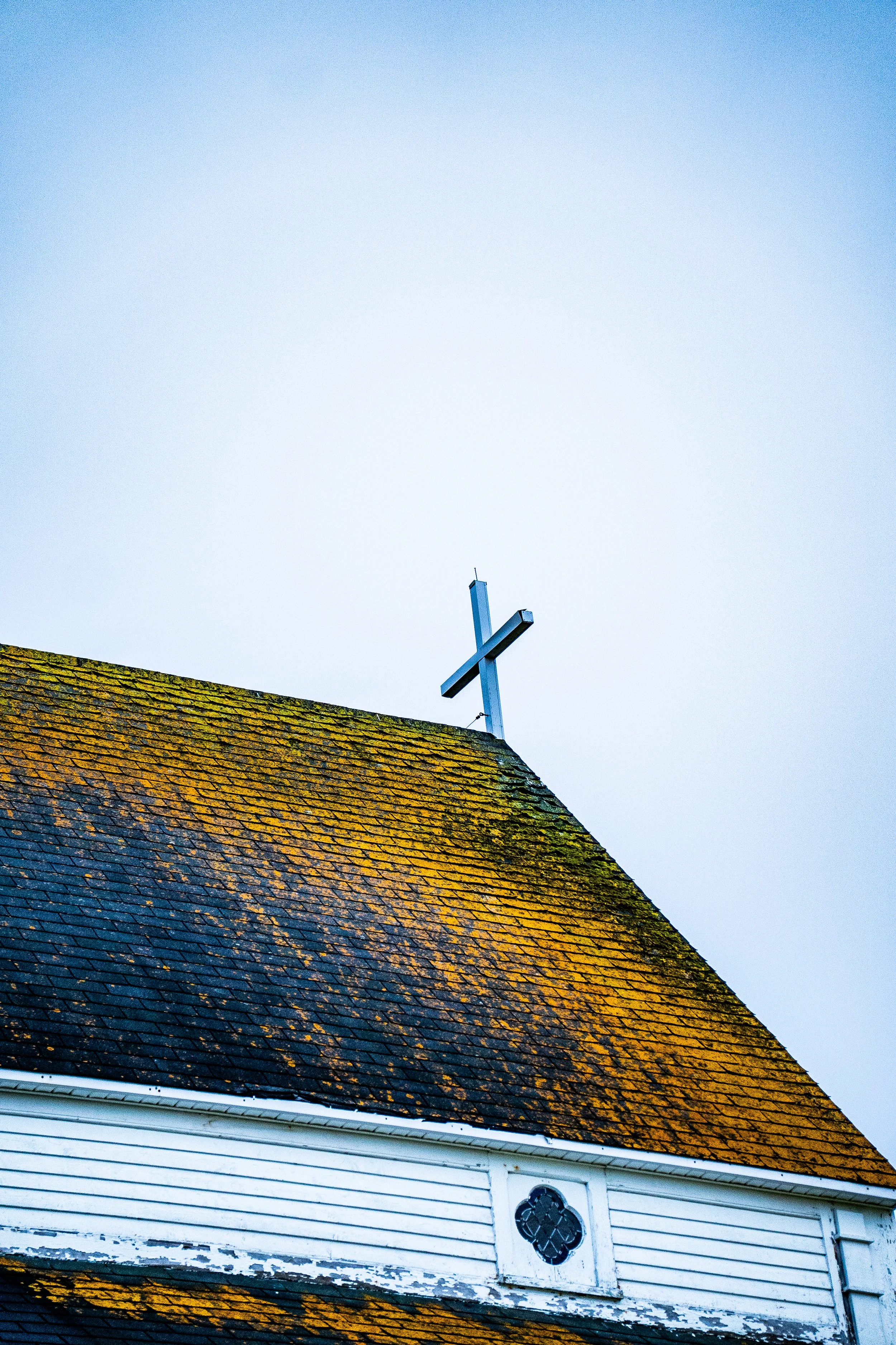 Close-up of church roof with a wooden cross on top, moss and weathered shingles, white clapboard siding below, under a clear sky.