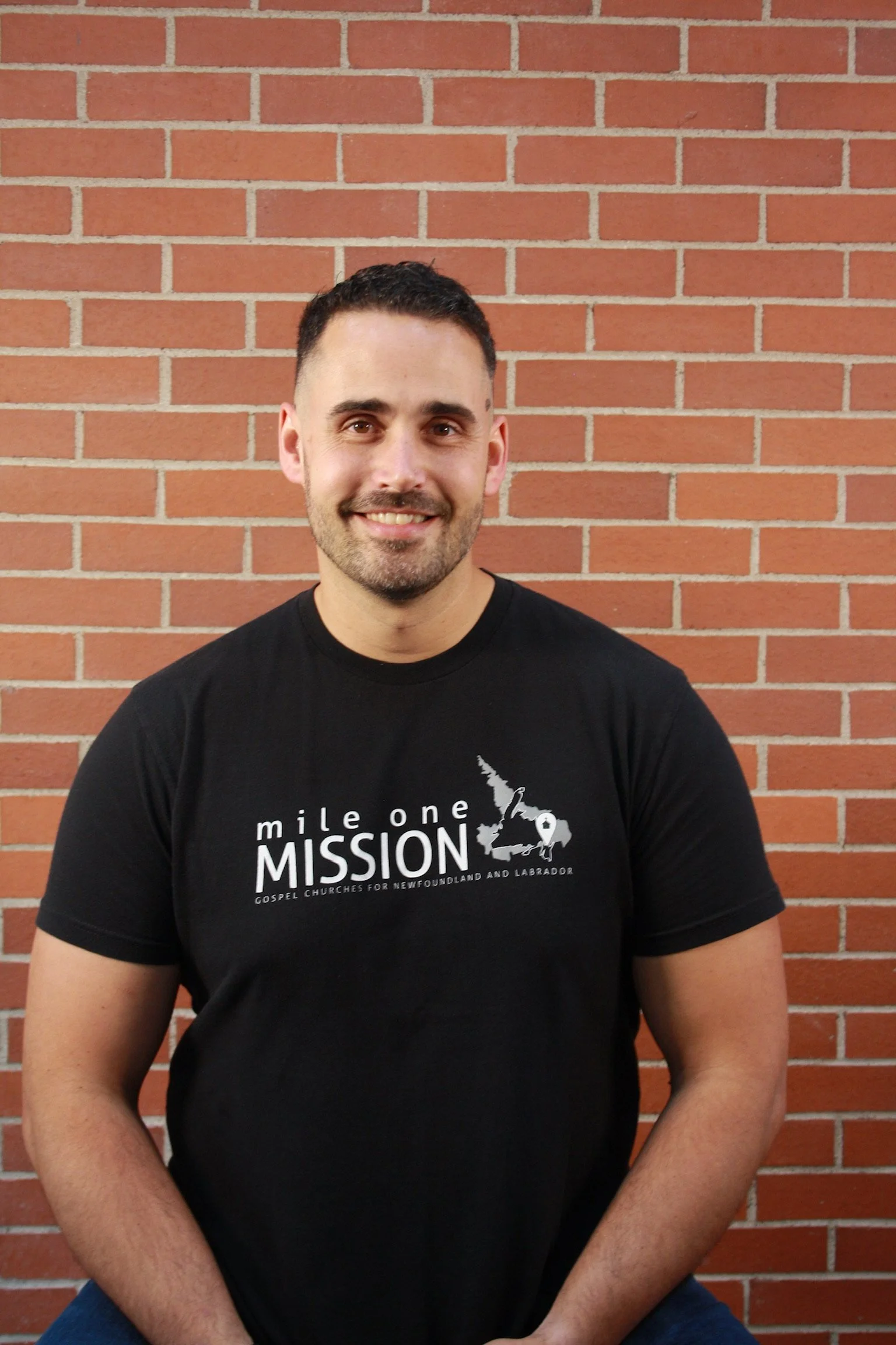 A smiling man with short dark hair and a beard, wearing a black t-shirt, sitting against a brick wall background.