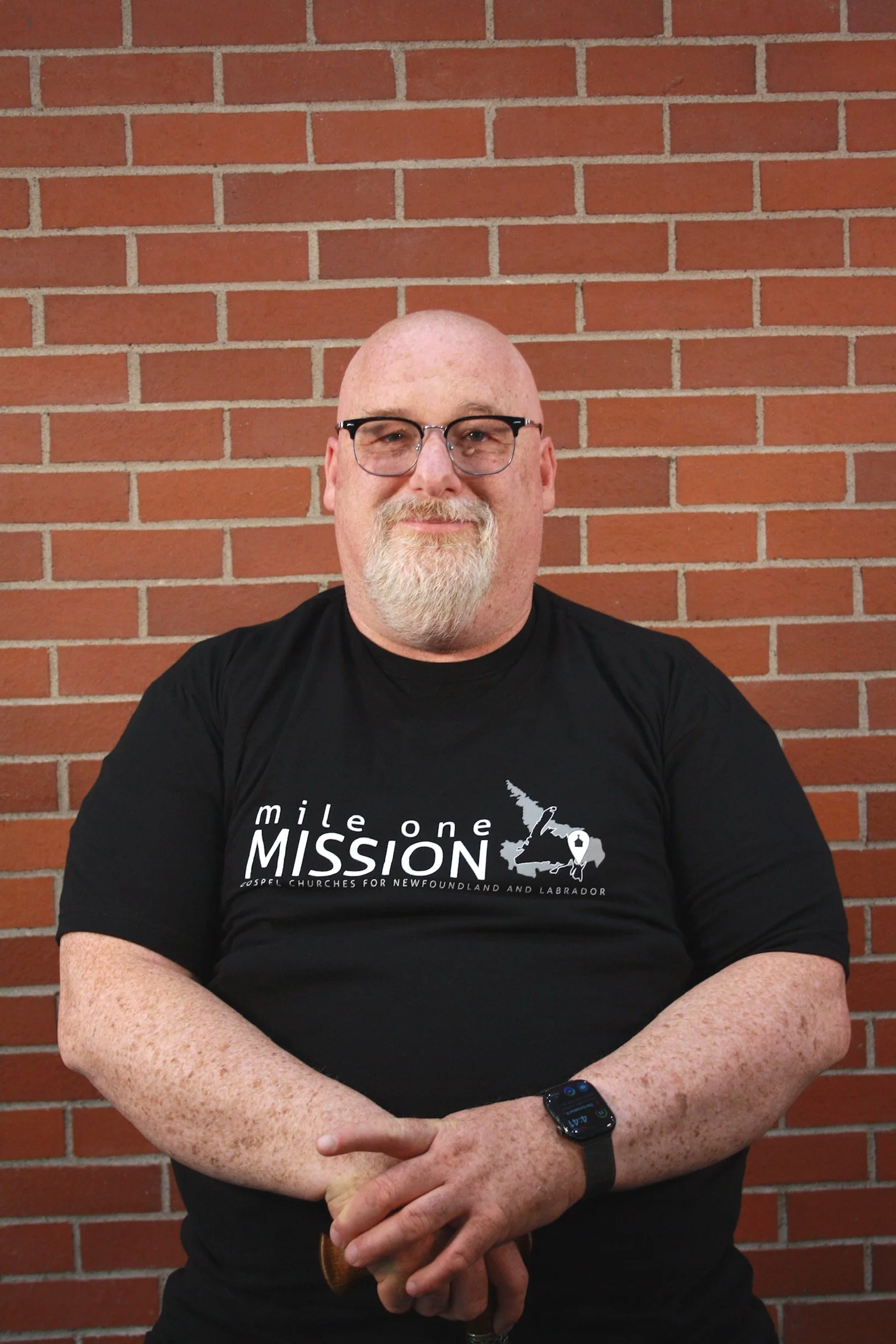 A man with glasses and a goatee, wearing a black t-shirt and smartwatch, is standing in front of a red brick wall.