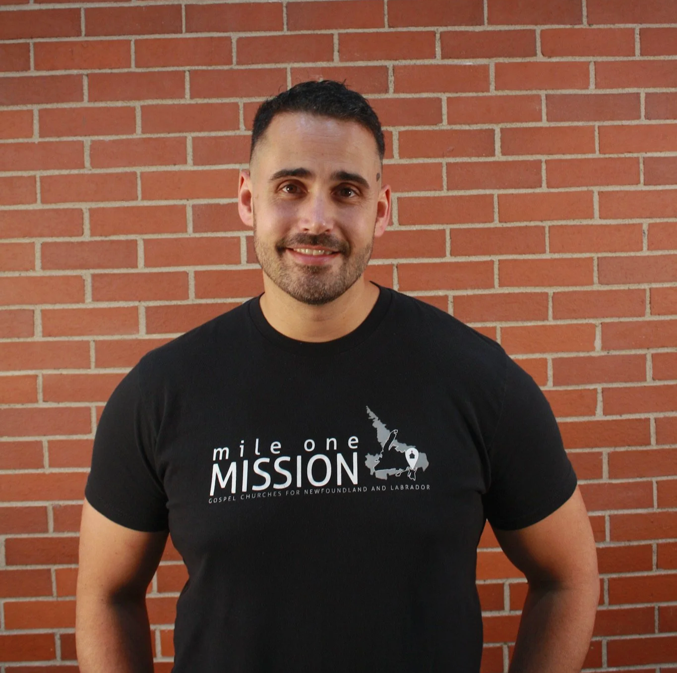 A man with short dark hair and a beard wearing a black t-shirt standing in front of a red brick wall.