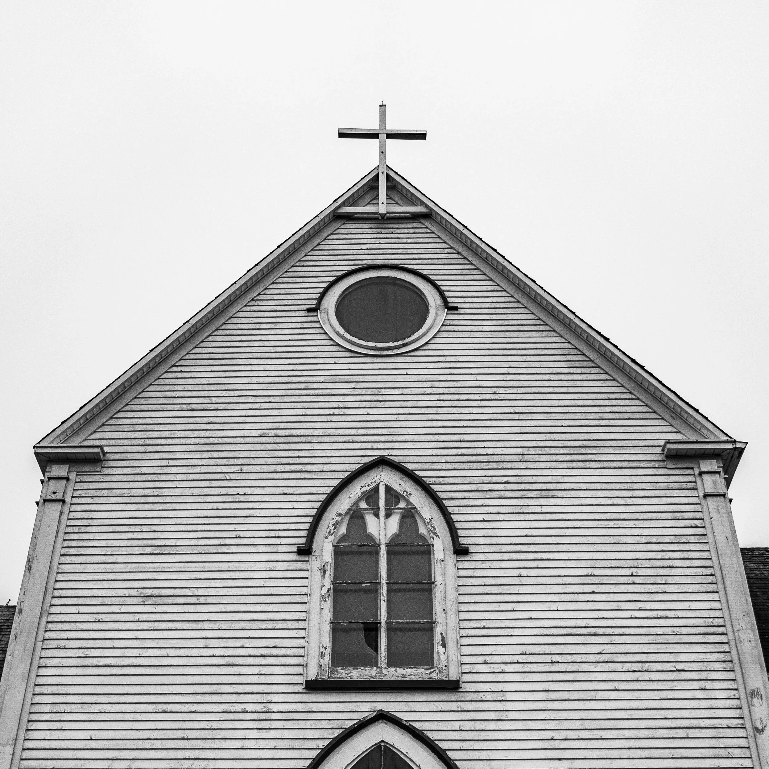 Black and white photo of the top part of a church with a cross on the steeple and arched windows, showing weathered wooden siding.