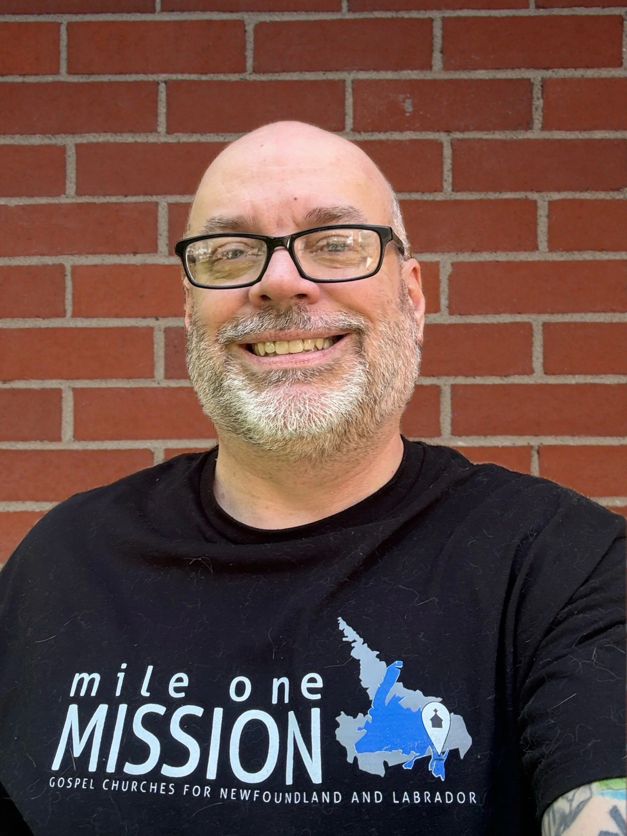 A man with glasses and a beard smiling in front of a brick wall, wearing a black T-shirt with a blue and white graphic and white text about missionary work in Newfoundland and Labrador.
