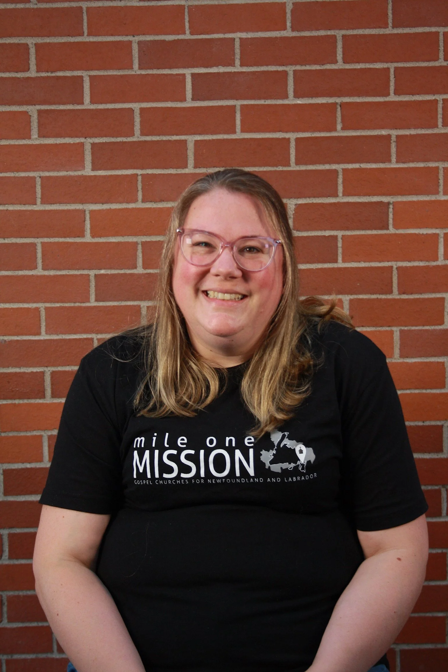 A woman with long blonde hair wearing pink glasses and a black T-shirt with white text, sitting against a red brick wall, smiling at the camera.