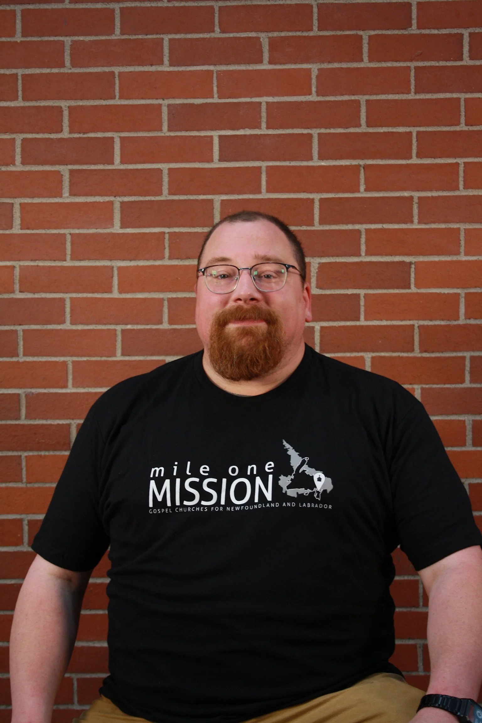 A man with glasses and a beard sitting against a red brick wall. He is wearing a black T-shirt with the text 'mile one MISSION Gospel Churches for Newfoundland and Labrador' and khaki pants.