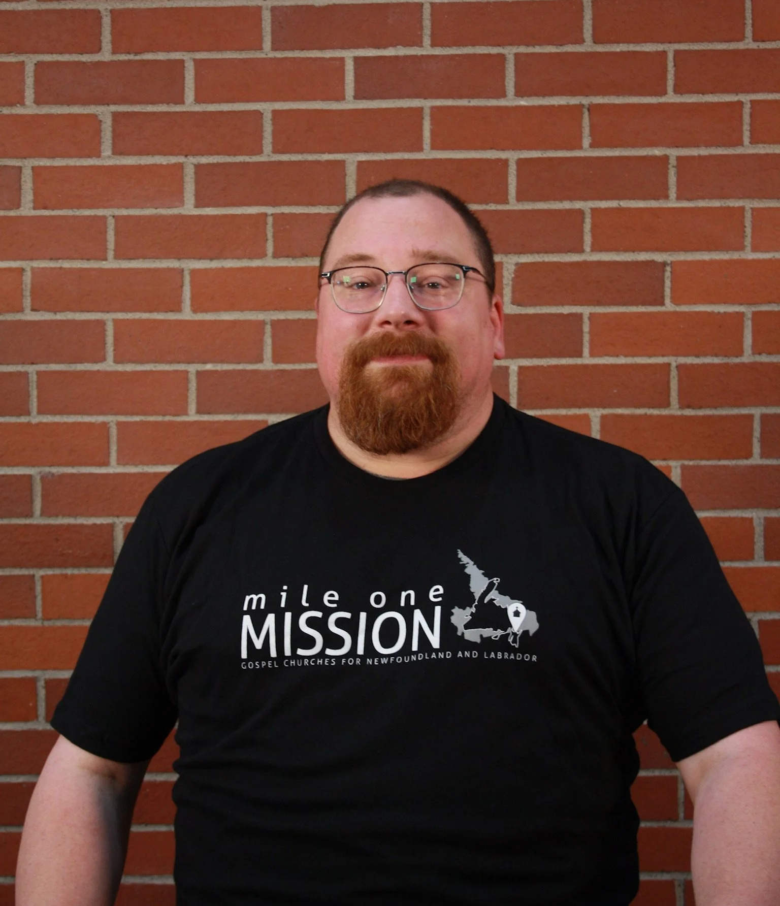 A man with glasses and a red beard wearing a black T-shirt with a map of Newfoundland and Labrador and the words 'mile one MISSION' stands against a red brick wall.